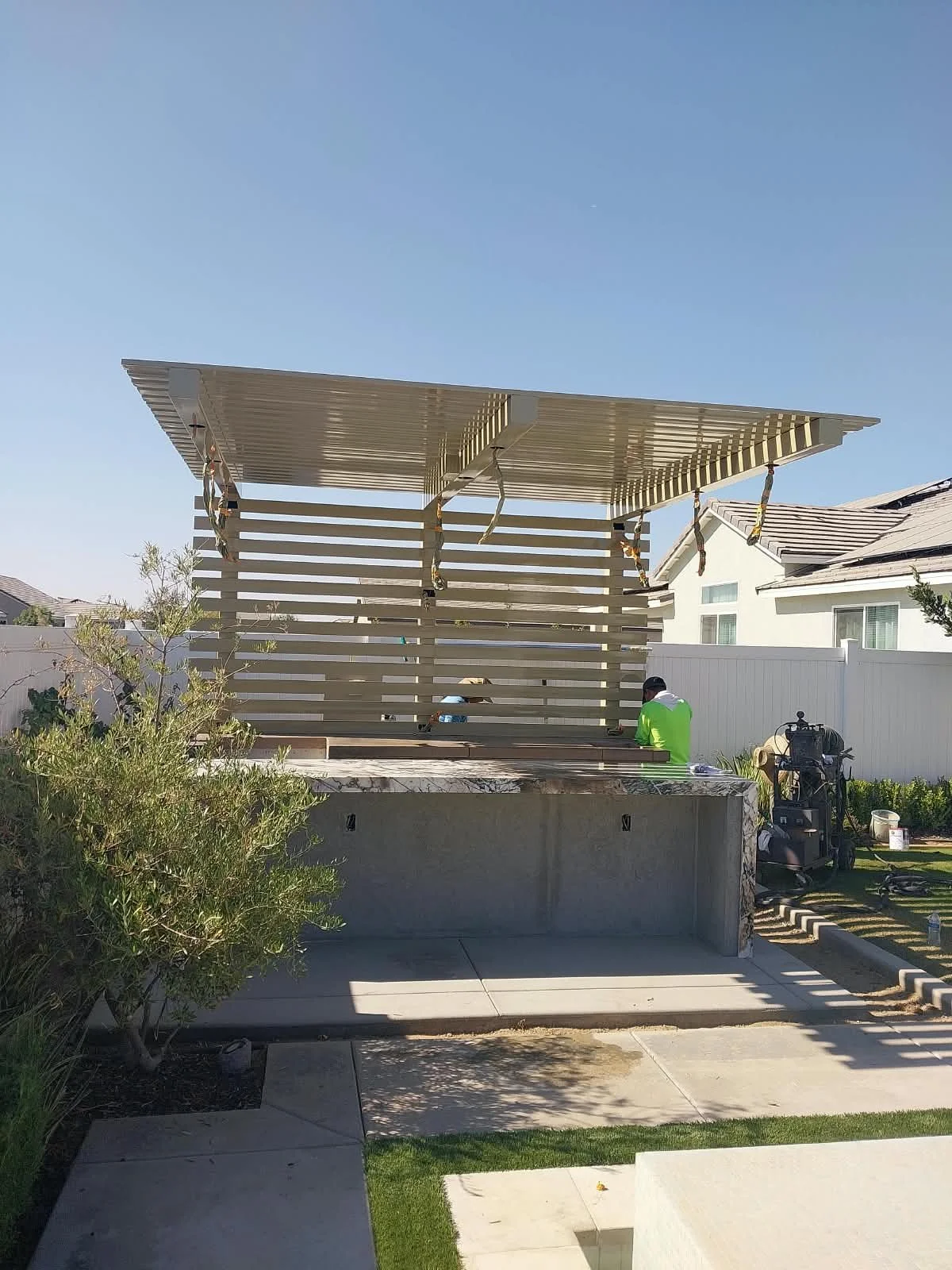A residential backyard with a modern pergola under construction. Two workers are seen, one wearing a hat and the other in a lime green jacket, working on the structure. There is construction equipment and materials nearby, with a white fence and neig
