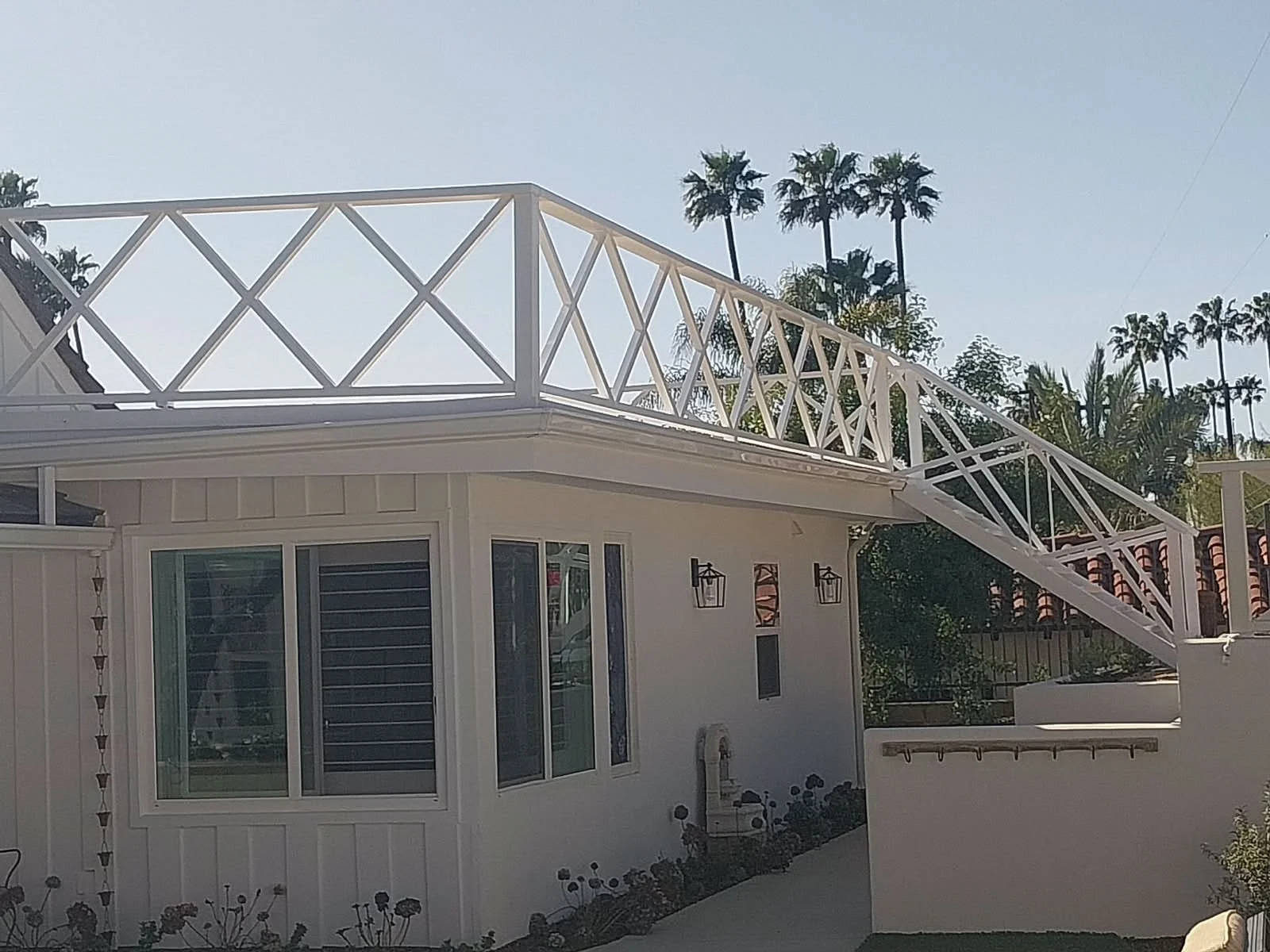 White house with a sliding glass door and a decorative outdoor light fixture, topped by a white railing and an elevated deck with palm trees in the background.