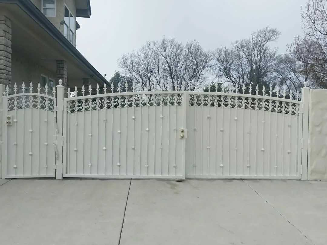 White decorative metal gate with curved top and pointed finials, installed on a concrete driveway in front of a house with stone and siding exterior, bare trees in the background.