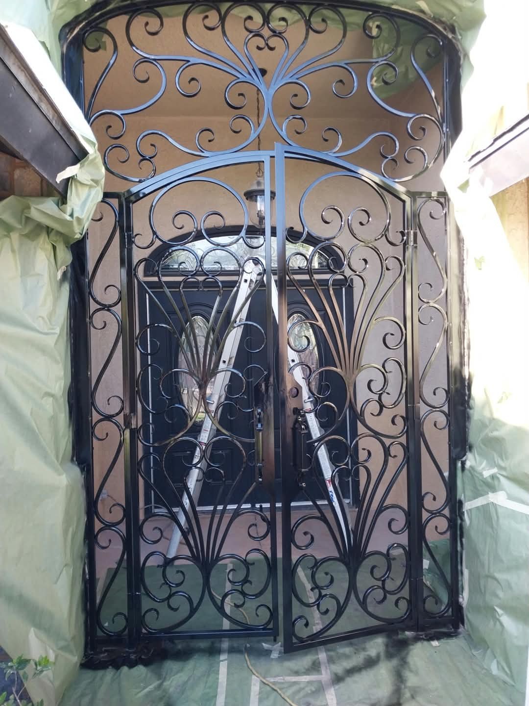 Black wrought iron decorative double front door with intricate scrollwork design, flanked by temporary protective yellow cloth coverings.