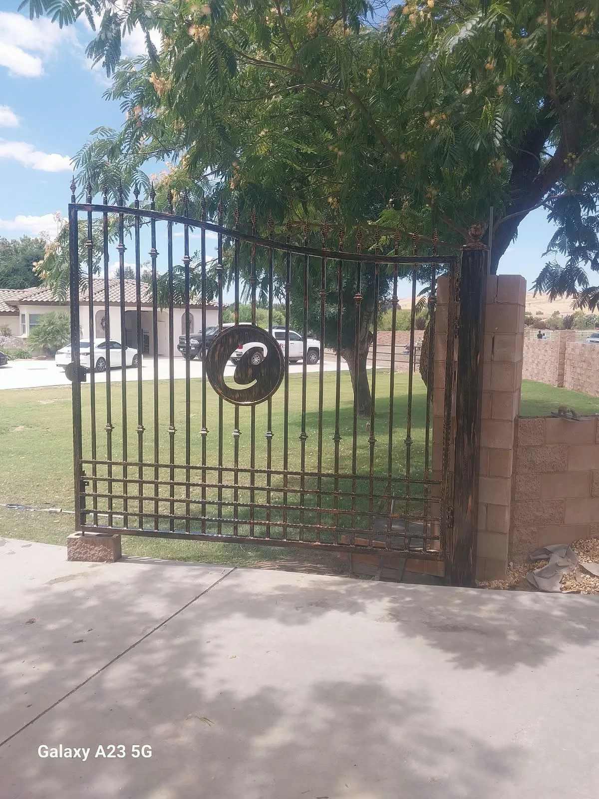 Close-up of an open iron gate with a decorative spiral design in the center, leading to a grassy yard with trees and a house in the background under a partly cloudy sky.