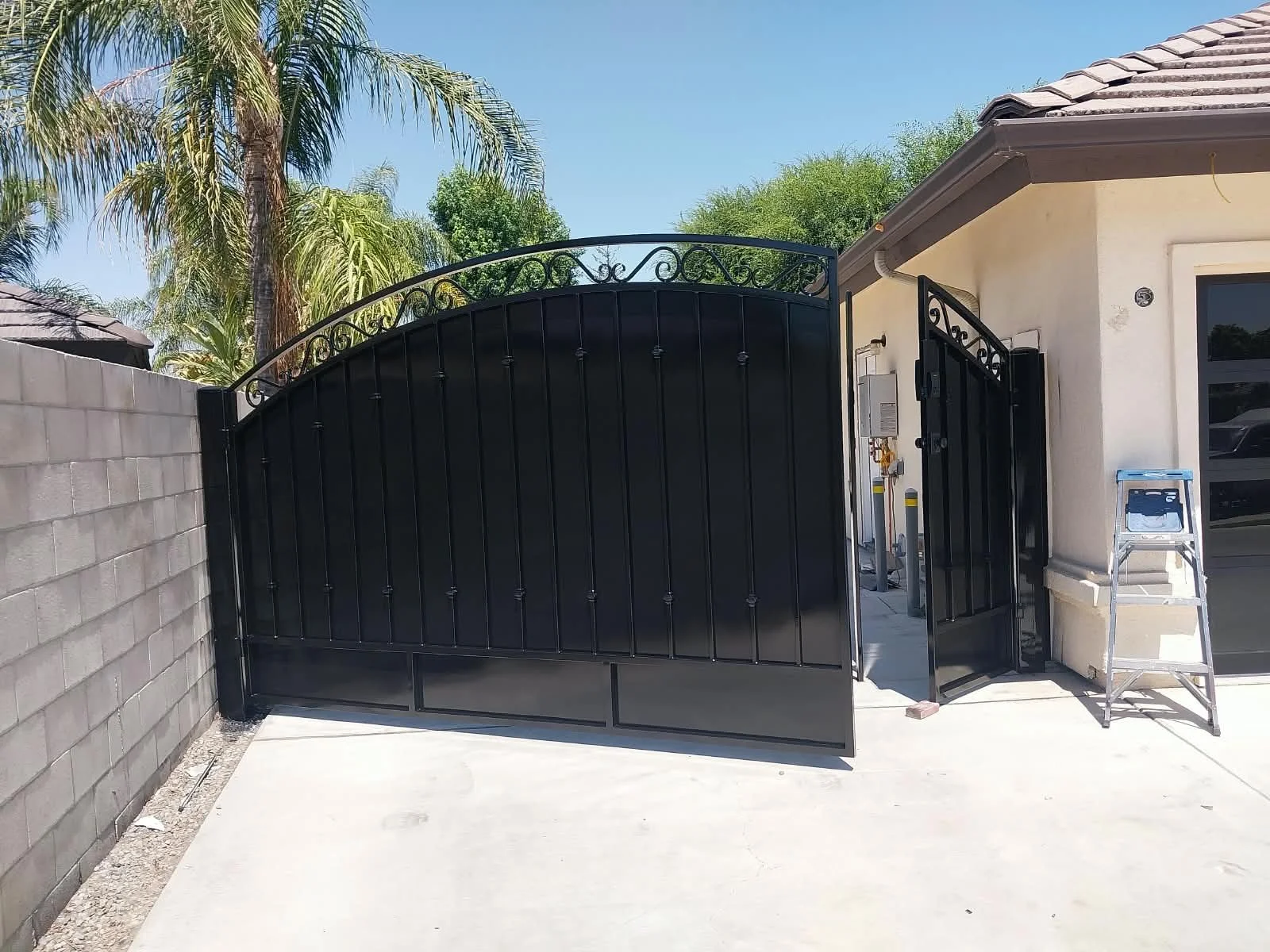 Black wrought iron gate partially open in front of a white house with a tiled roof, to the right of the gate is a ladder, and on the left is a gray brick wall, with palm trees and greenery in the background and a clear blue sky.