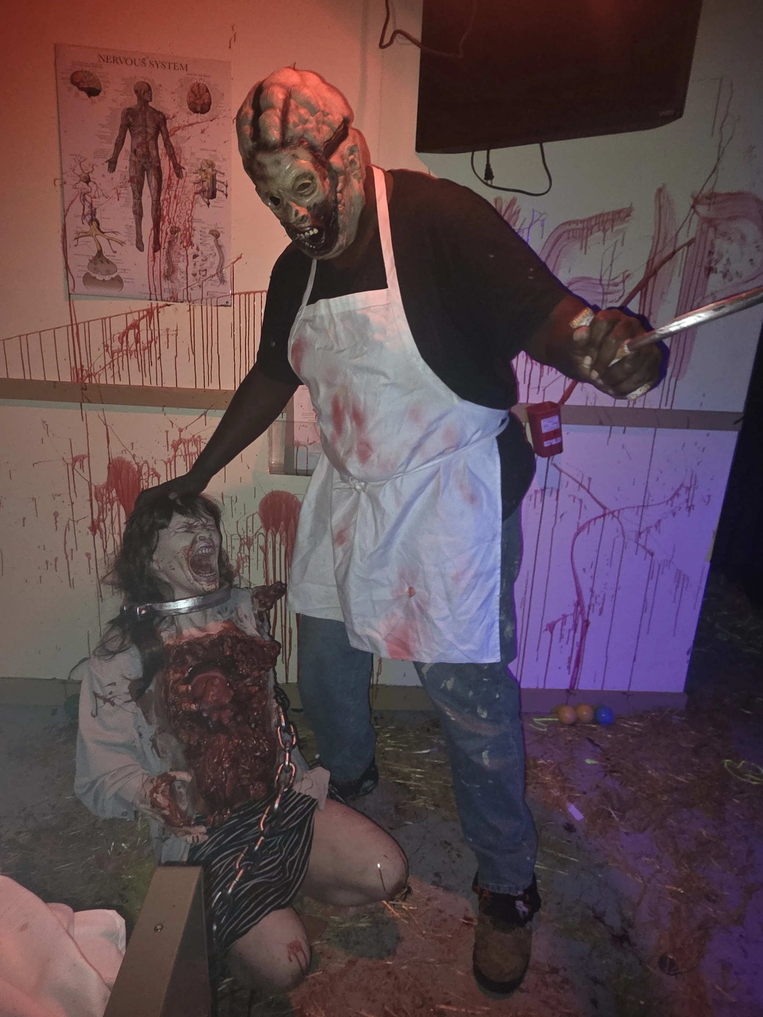 Halloween Manor Detroit actor wearing a horror mask and blood-stained apron beside a chained zombie character inside the haunted attraction.