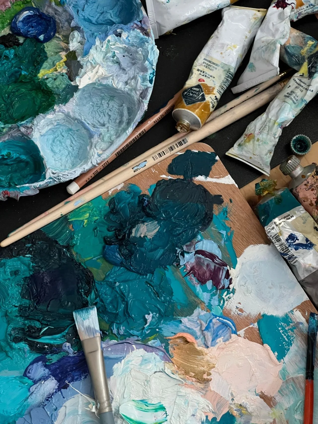 Messy artist's workspace with various tubes of paint, paintbrushes, a palette, and mixed paint on a wooden surface.