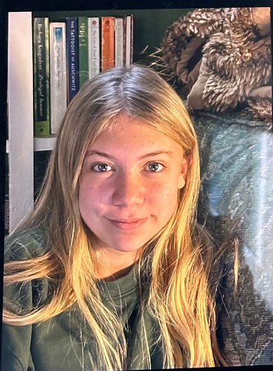 A young girl with long blonde hair and green eyes looking at the camera, sitting in front of a bookshelf and a plush gray blanket.