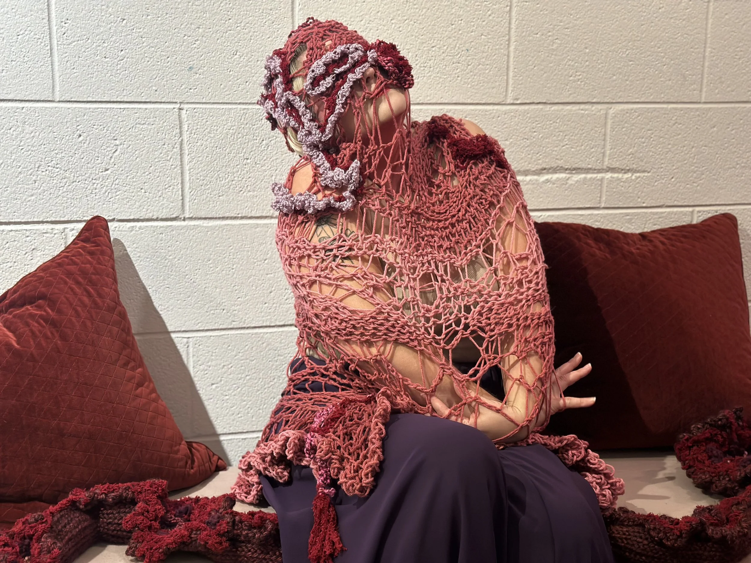 Person sitting on the floor against a white brick wall, surrounded by red pillows and a red blanket, wearing a pink crochet dress, with a crocheted mask covering their face.