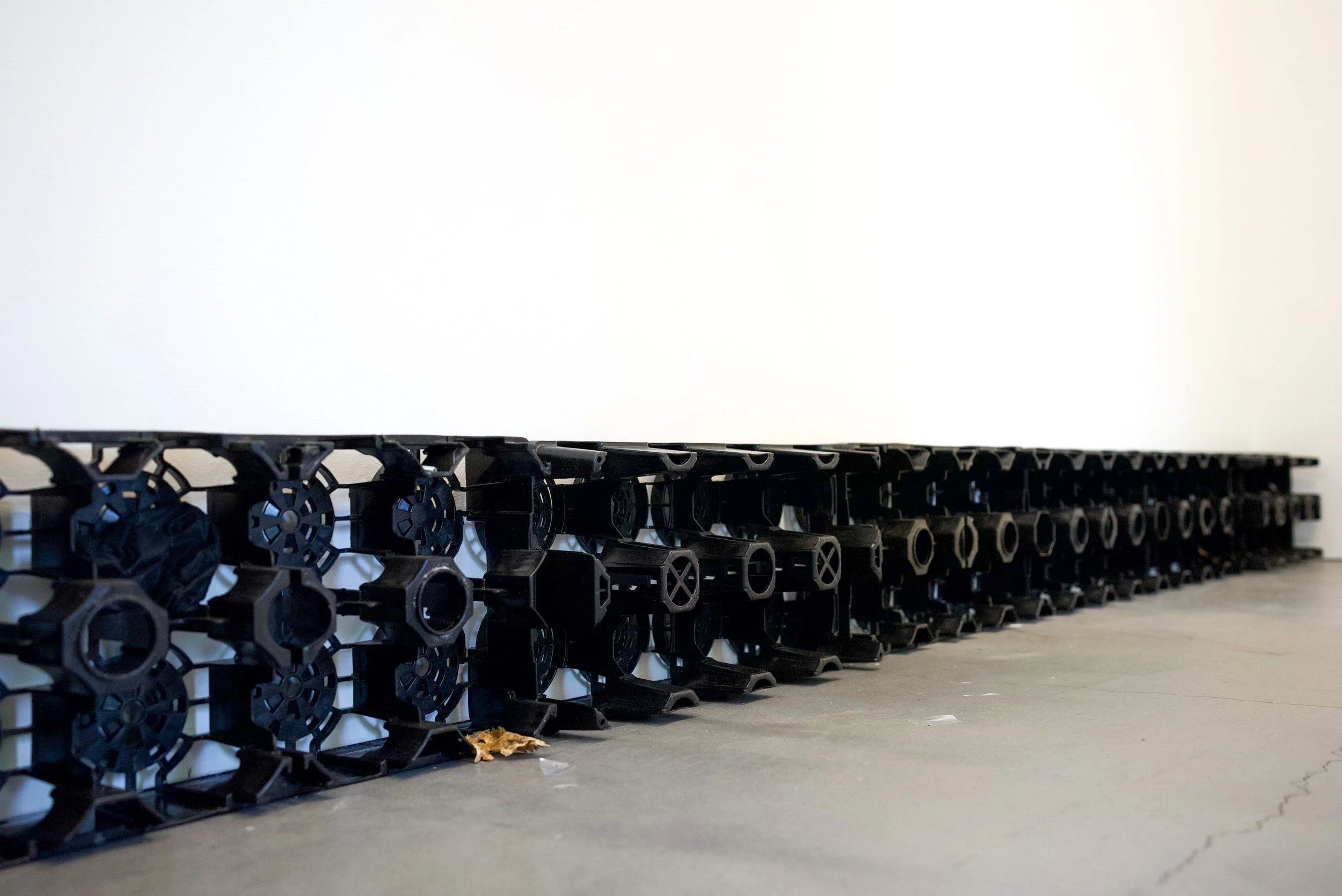 Stack of black plastic shipping crates for soda bottles placed against a white wall on a gray concrete floor, with some debris and a few leaves on the ground.