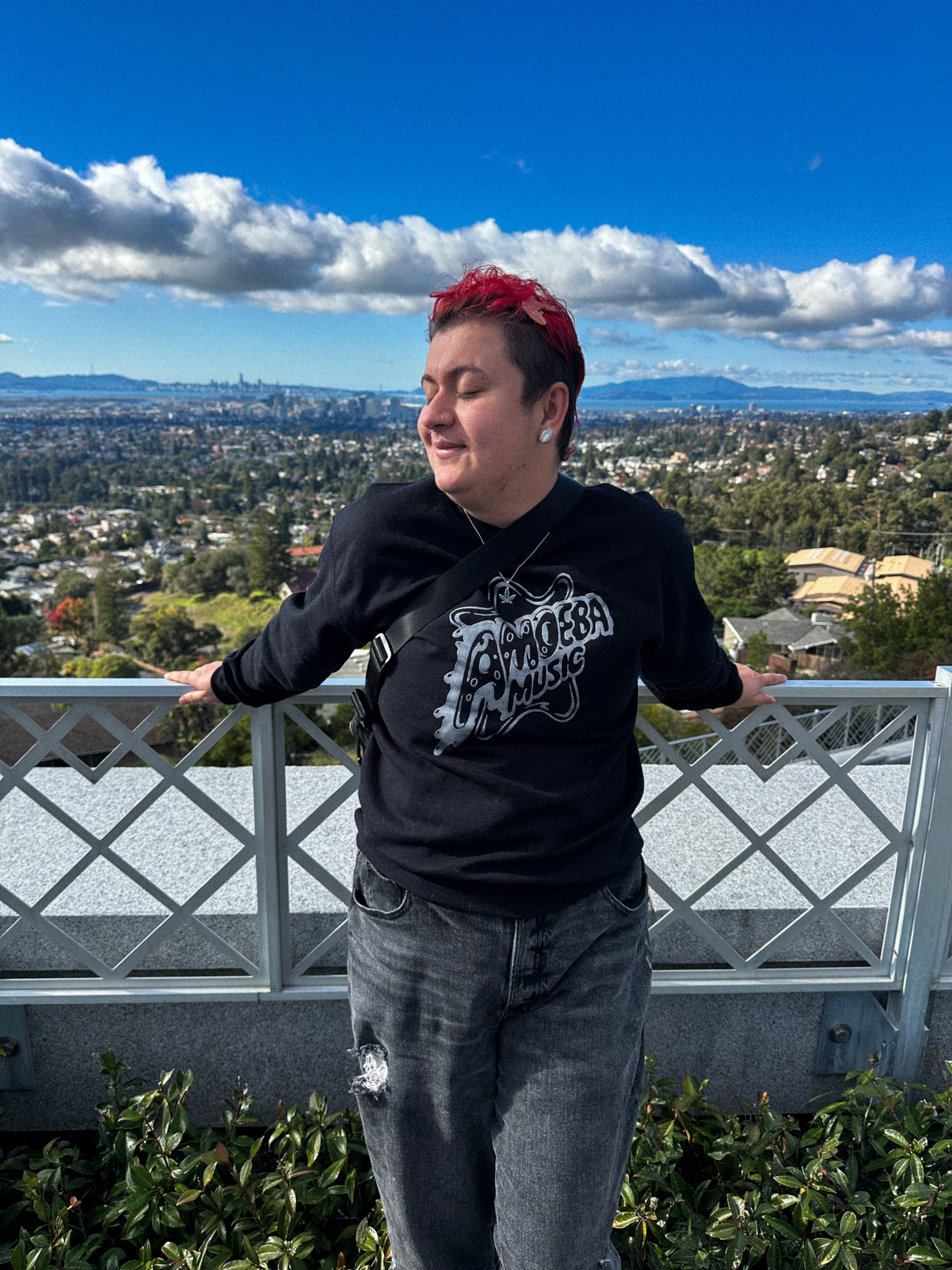 Person with short red hair and piercings standing outdoors on a balcony with cityscape and mountains in the background, wearing a black sweatshirt and ripped jeans.