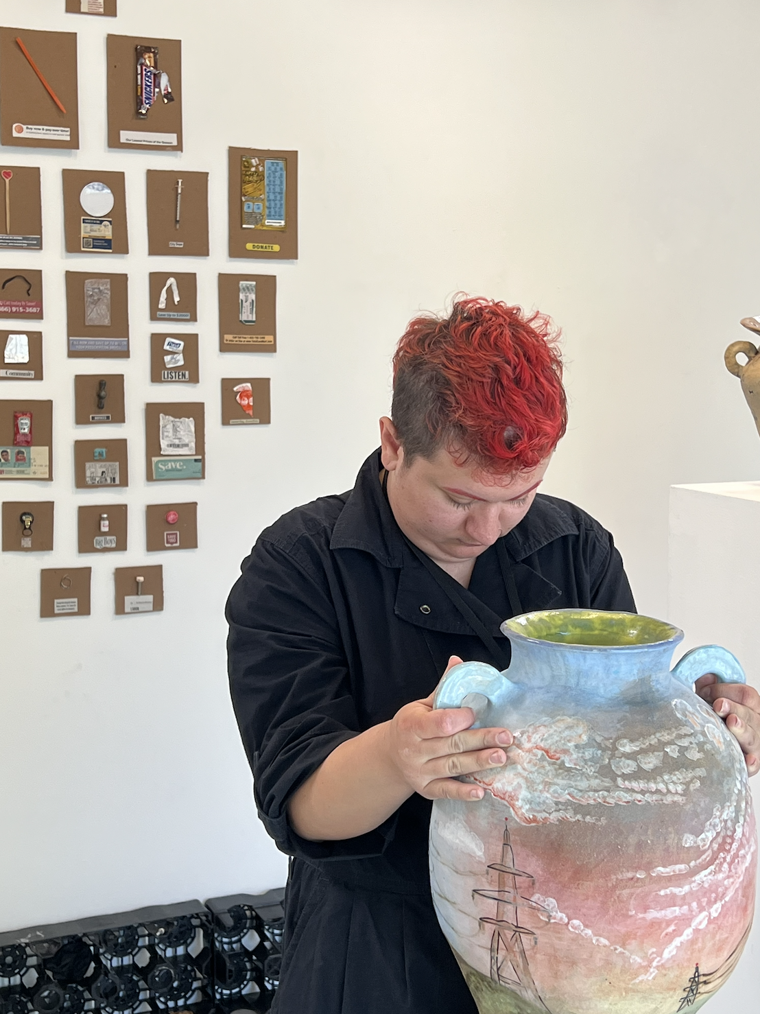 Person with red hair inspecting a large decorative ceramic vase with painted landscape and power lines.