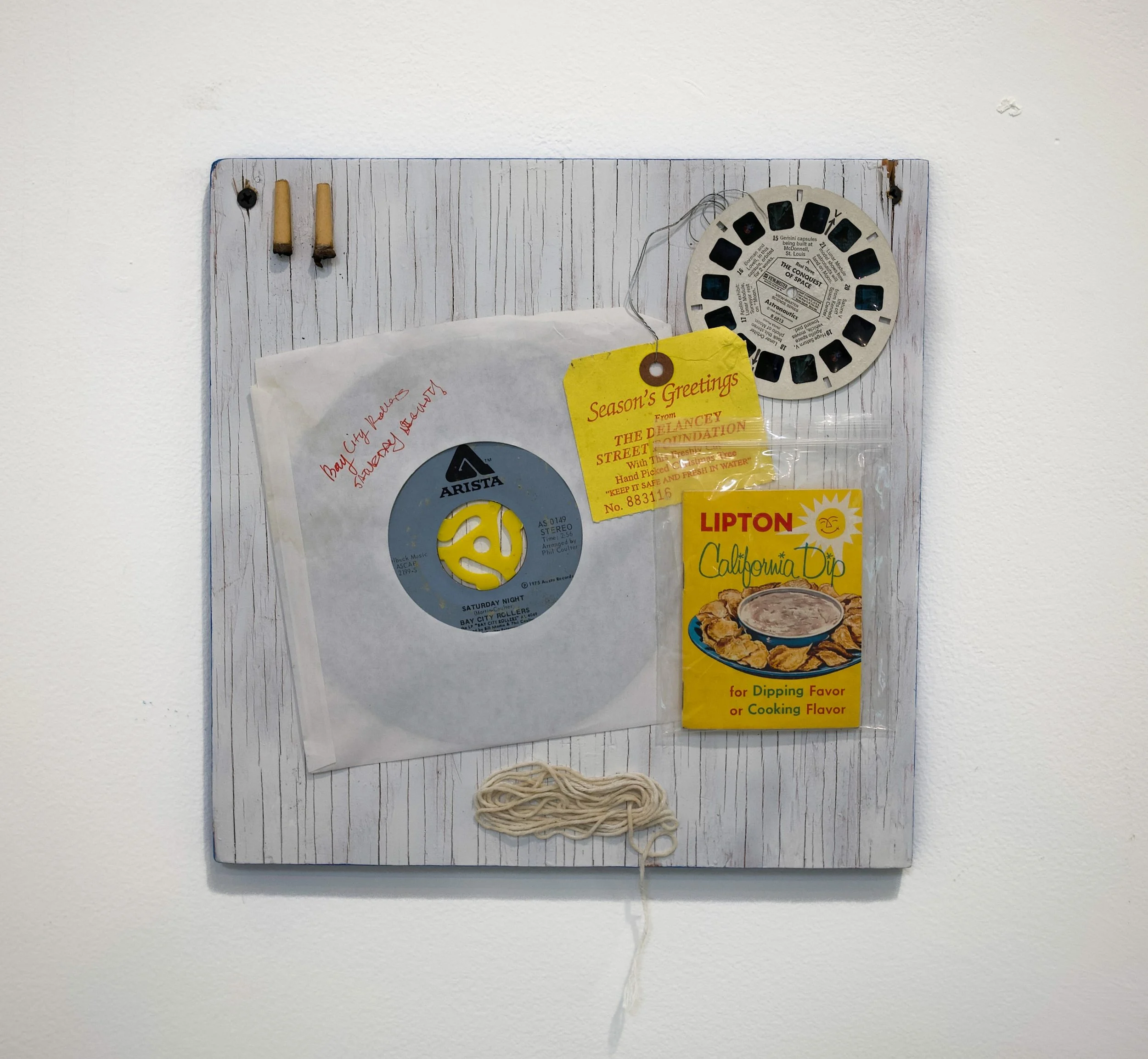 A wall-mounted wooden board with found objects such as a 45 record, string, and old recipe booklet collaged to make a repepitive composition.
