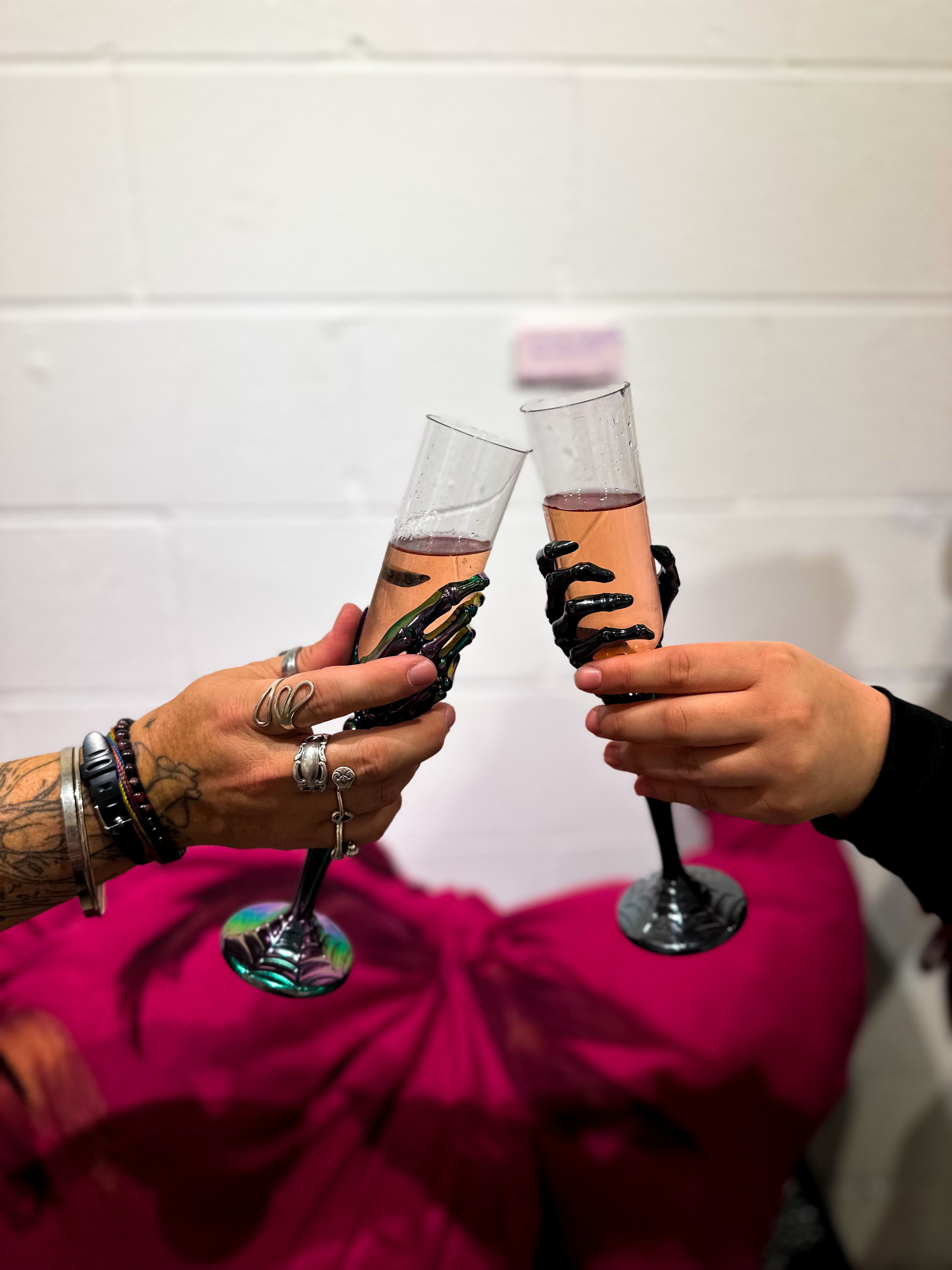Two hands, one with multiple rings and bracelets, the other with a wristband, holding glasses of pink wine in front of a white wall and pink fabric.