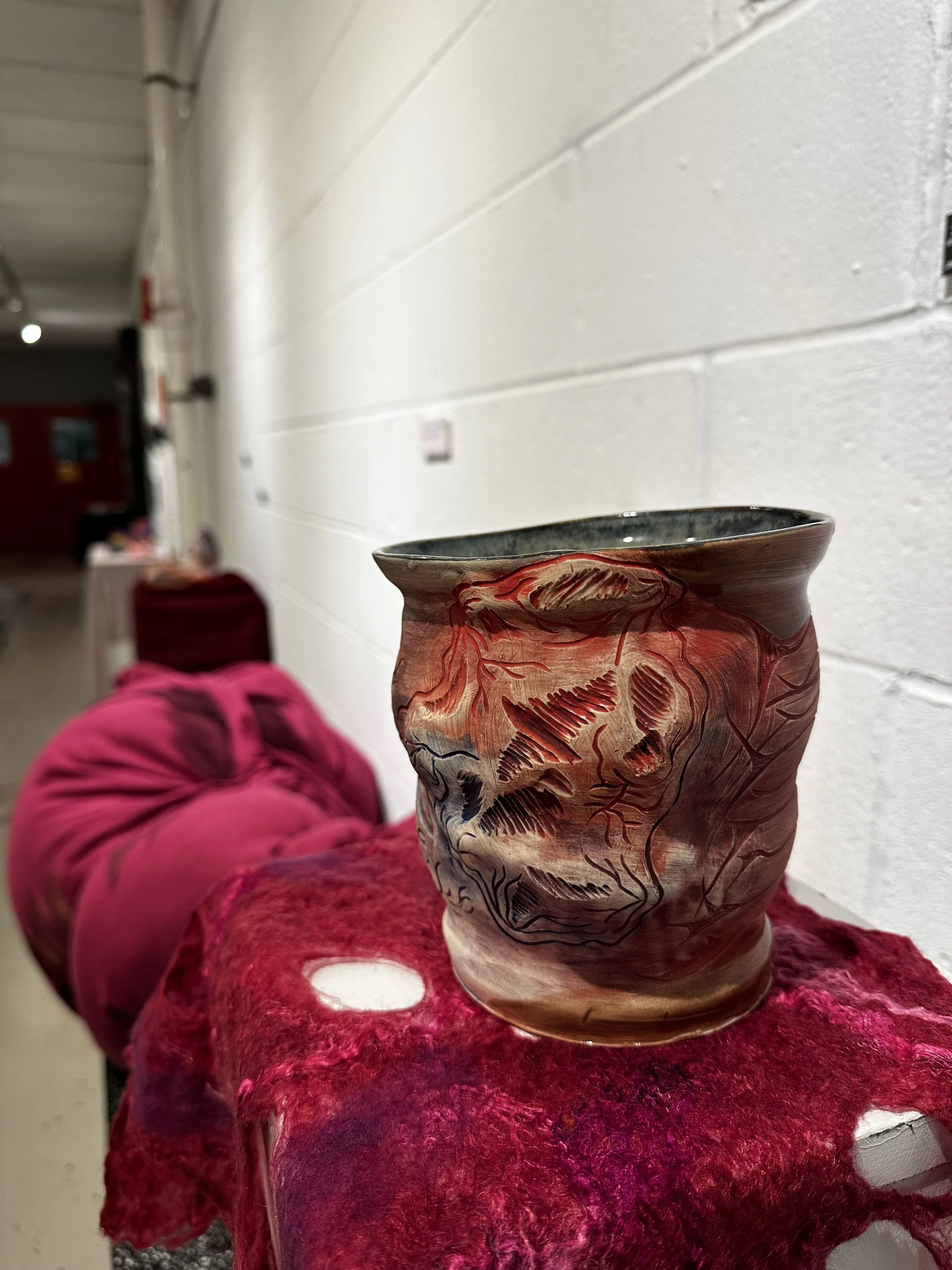 A decorative ceramic vase with a red and black organic design. The vase is resting on a red textured fabric surface. The background shows a white brick wall and a corridor.