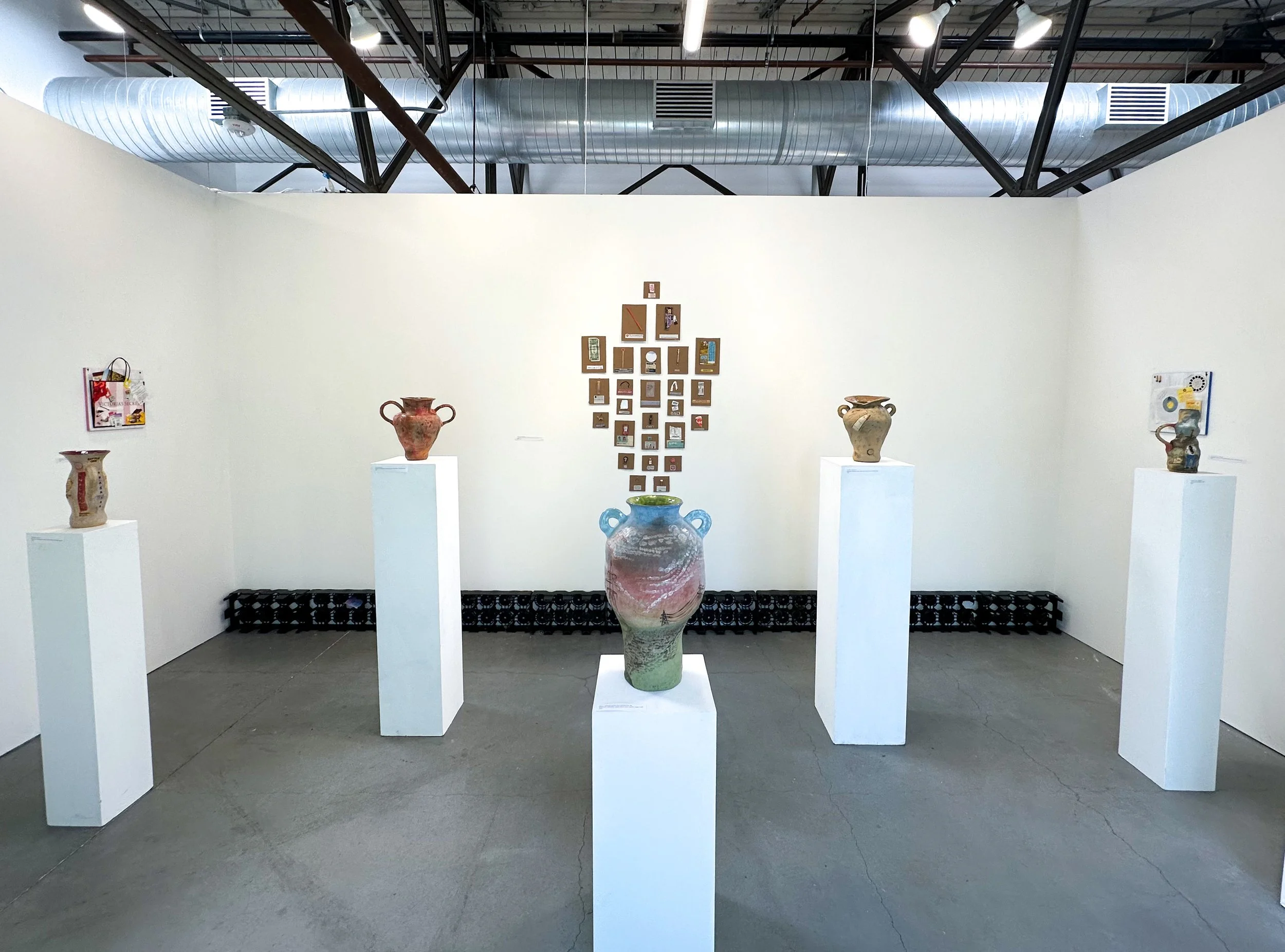 Art gallery exhibit displaying ancient pottery pieces on white pedestals, with a wall featuring small framed artworks and text in the background. Large ventilation ductwork and industrial ceiling structure are visible.