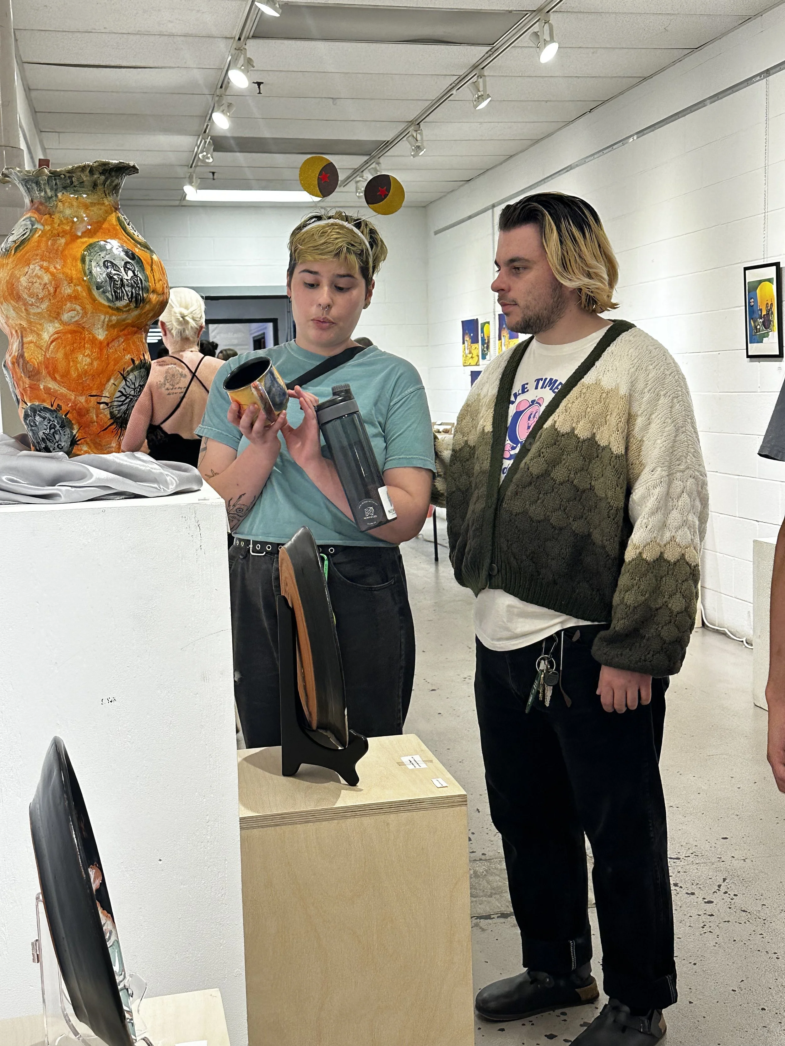 Two young adults, a woman with short blonde hair and a man with long blonde hair, are looking at a small mug the woman is holding in an art gallery. The woman wears a teal T-shirt and has tattoos on her right arm, while the man wears a beige and gree