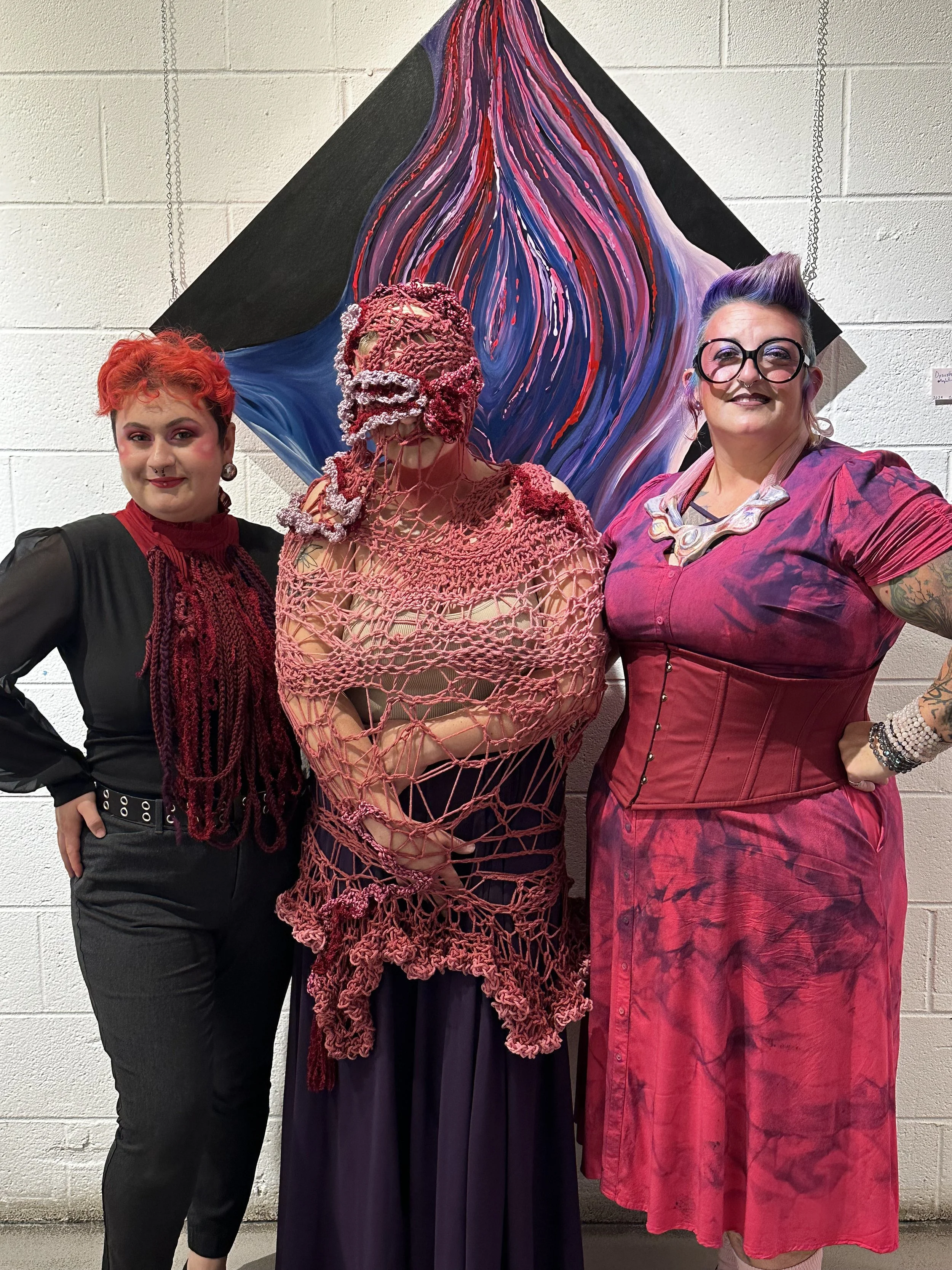 Three people dressed in artistic and colorful clothing standing in front of a large, abstract painting with dripped paint in shades of purple, red, and blue, against a white brick wall.