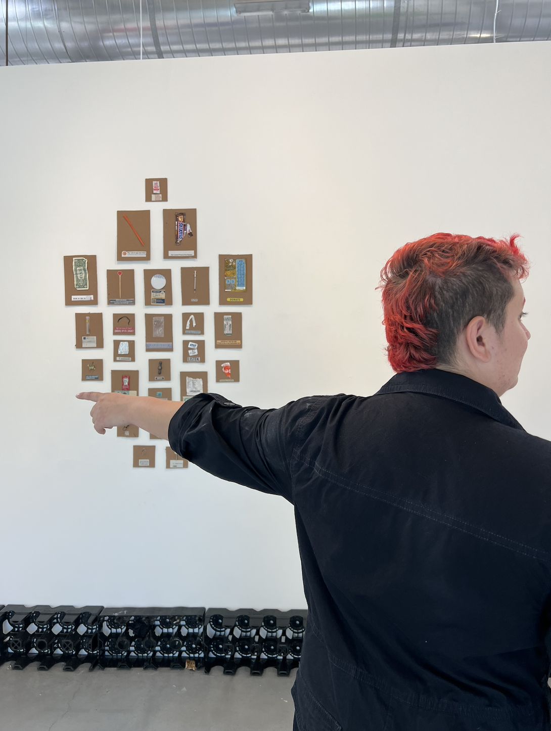 Person with short red hair pointing at a wall display of small art pieces of mixed objects arranged in a grid, with bottle-shipping crates lining the floor below.