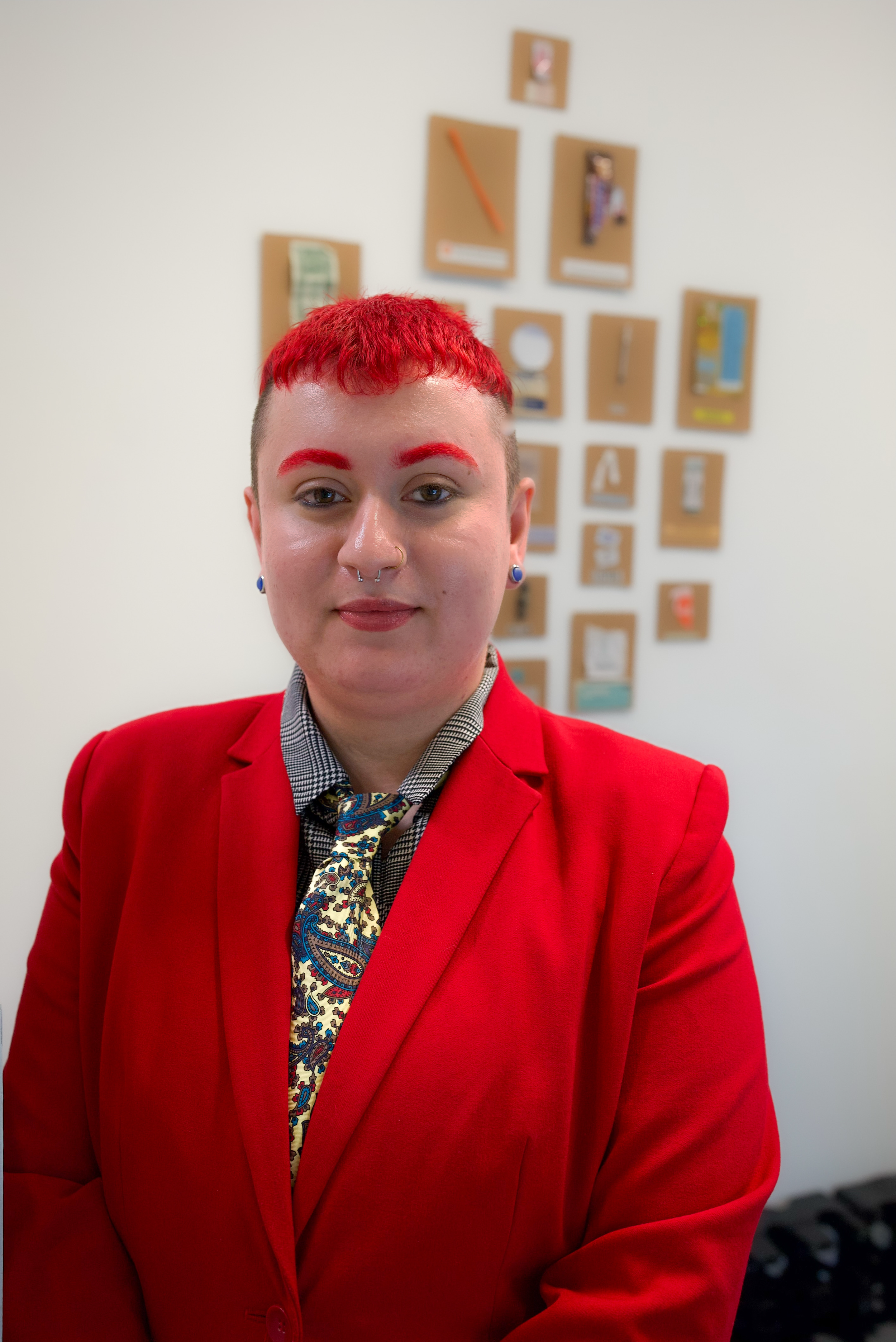 A person with short red hair and red eyebrows wearing a red blazer, a patterned shirt, and a colorful paisley tie, standing in front of a white wall with various pieces of art and photographs.