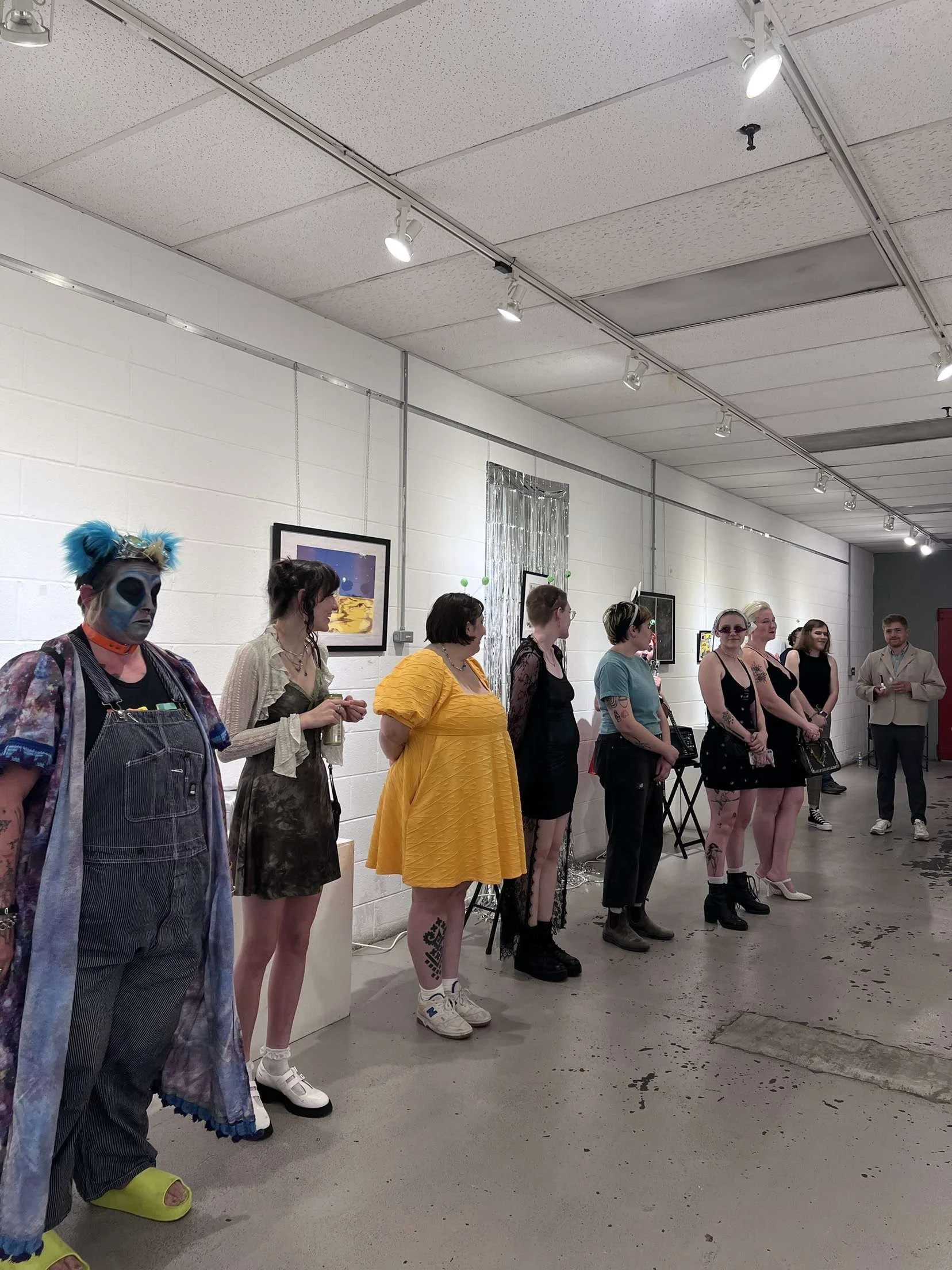 A group of people standing in a line in an art gallery, dressed in various costumes and outfits, with a white brick wall and artwork in the background.