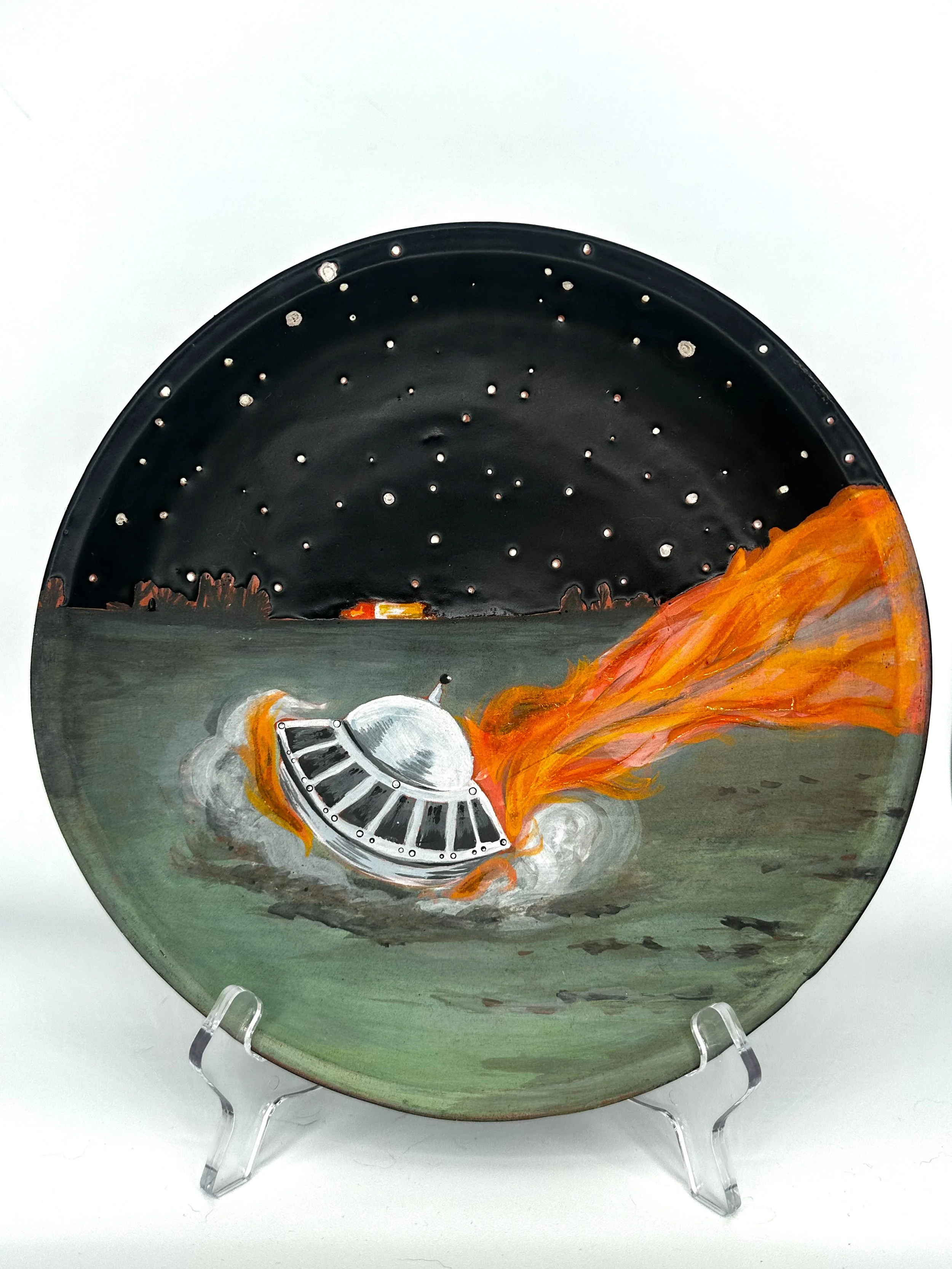 Painting on a ceramic plate of a UFO flying saucer crash-landing into a field at night time. 