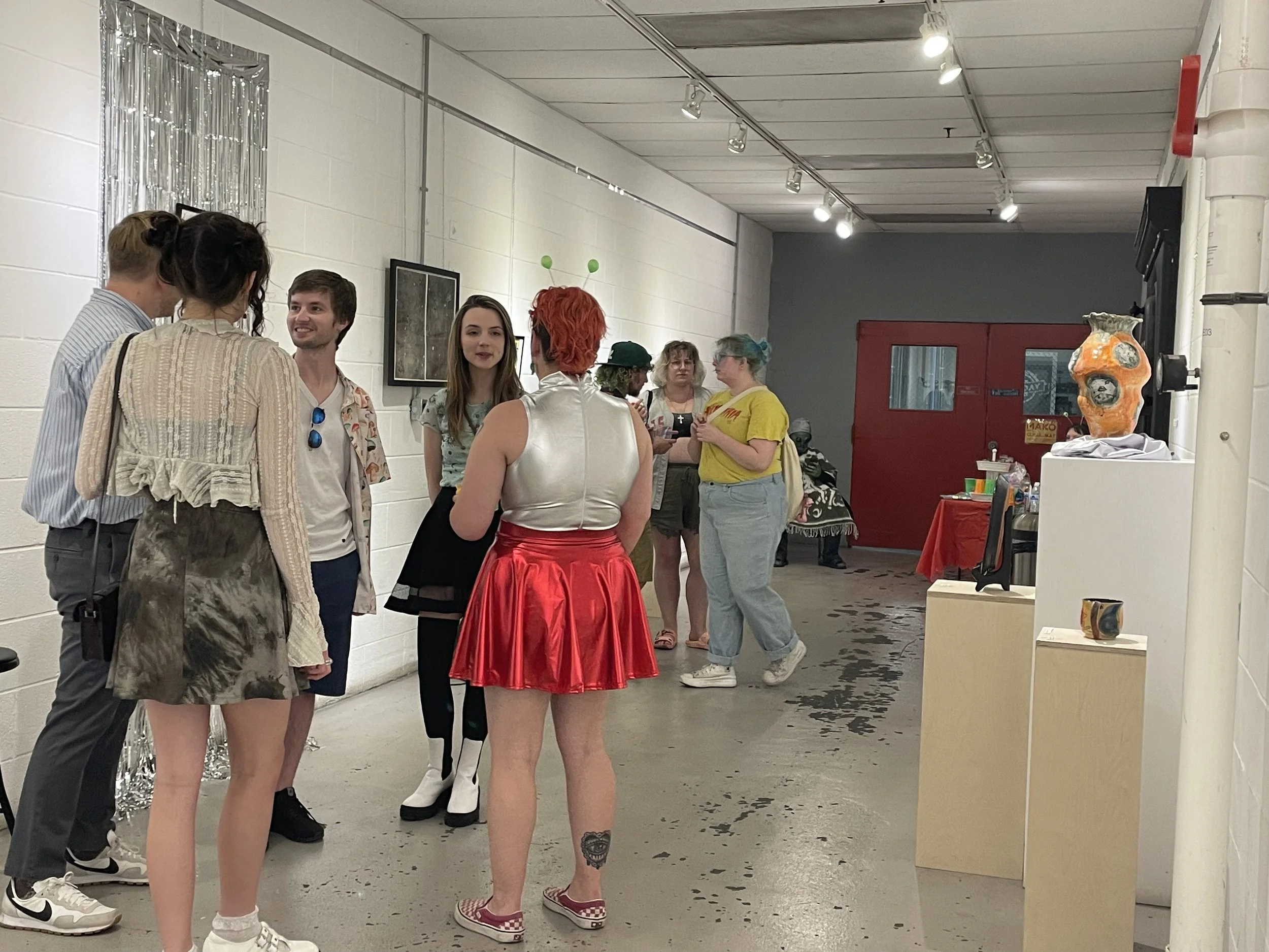A group of people standing and talking in an art gallery. Some of them are dressed in colorful, avant-garde clothing, including a person in a shiny white top with a red skirt and another with antennae headbands.