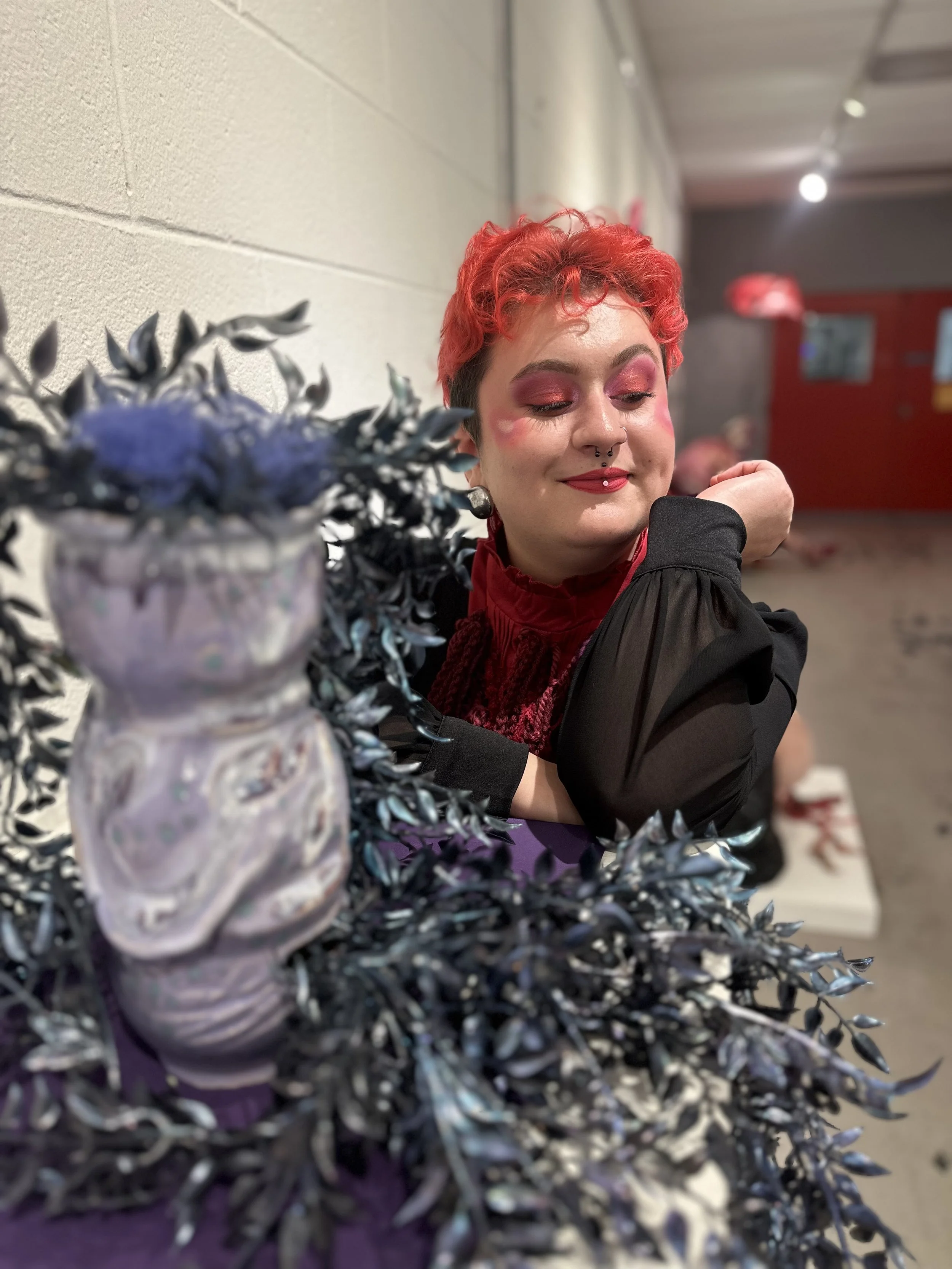 Person with short, curly red hair and makeup, smiling with eyes closed, sitting at a table with a decorative ceramic vase in the foreground.