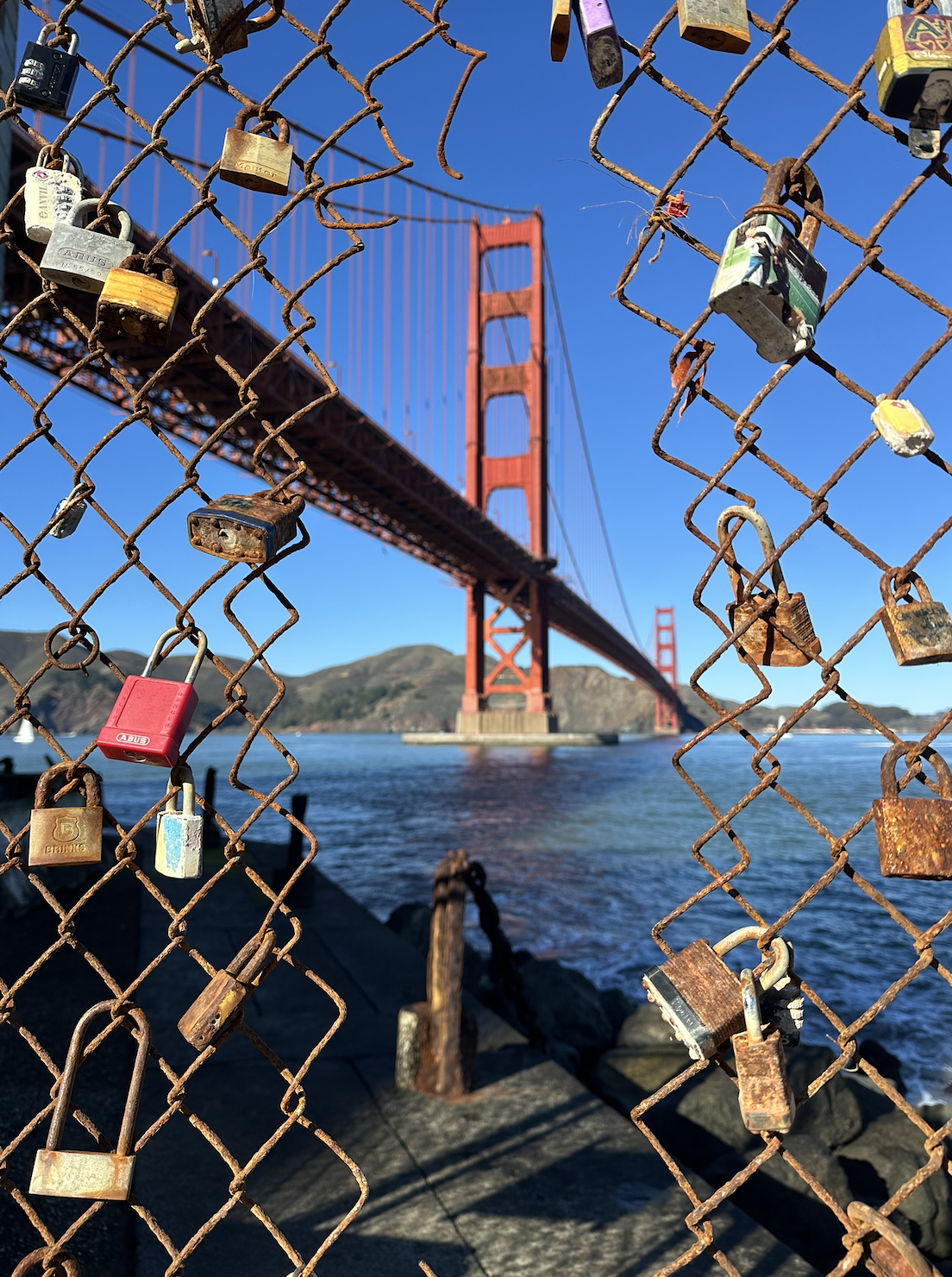 A rusty chain-link fence with numerous love locks attached to it, overlooking the Golden Gate Bridge in San Francisco under a clear blue sky.