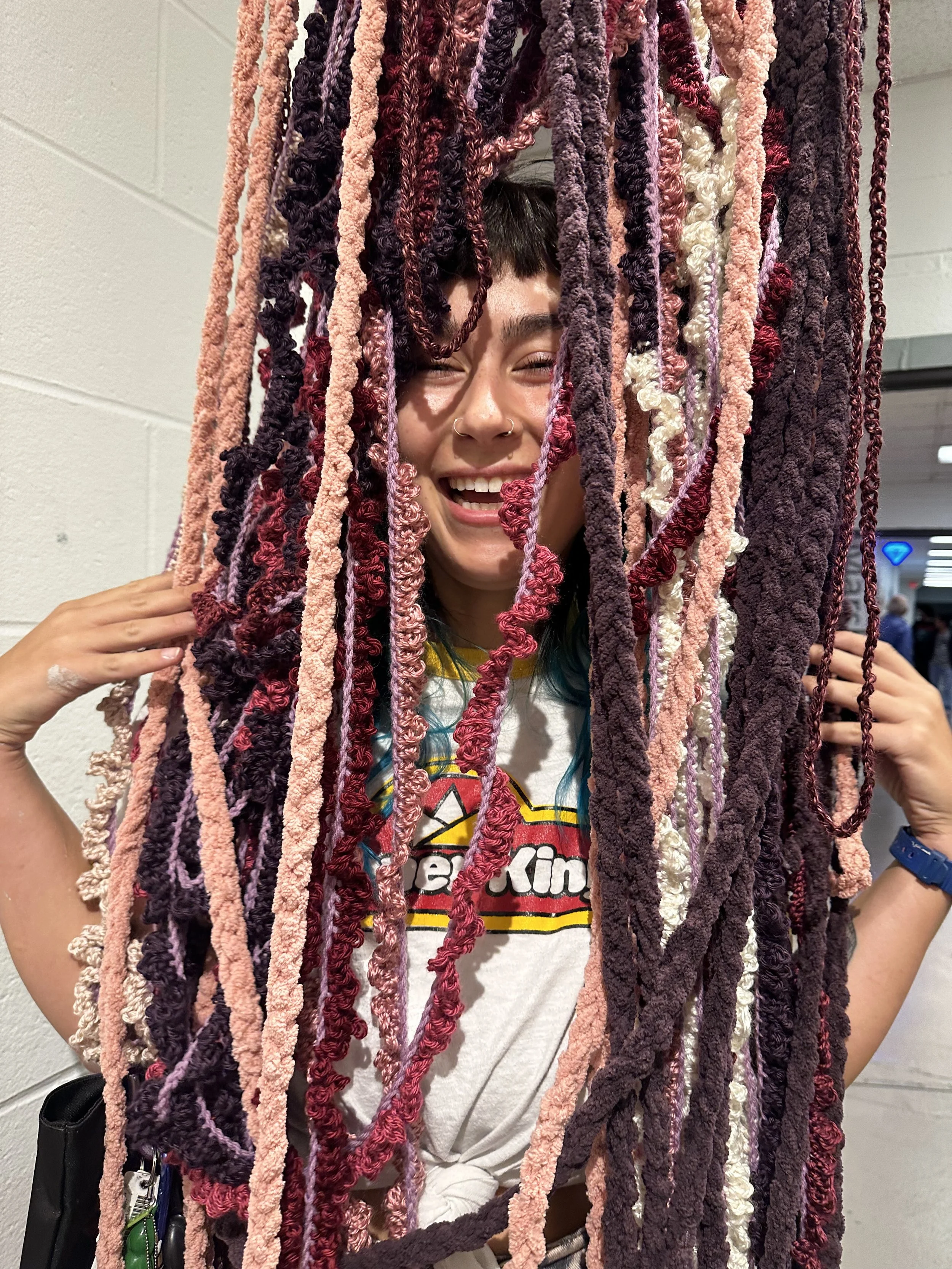 A person with colorful hair and a nose ring smiling behind hanging yarn strands in various shades of pink, purple, red, and white.