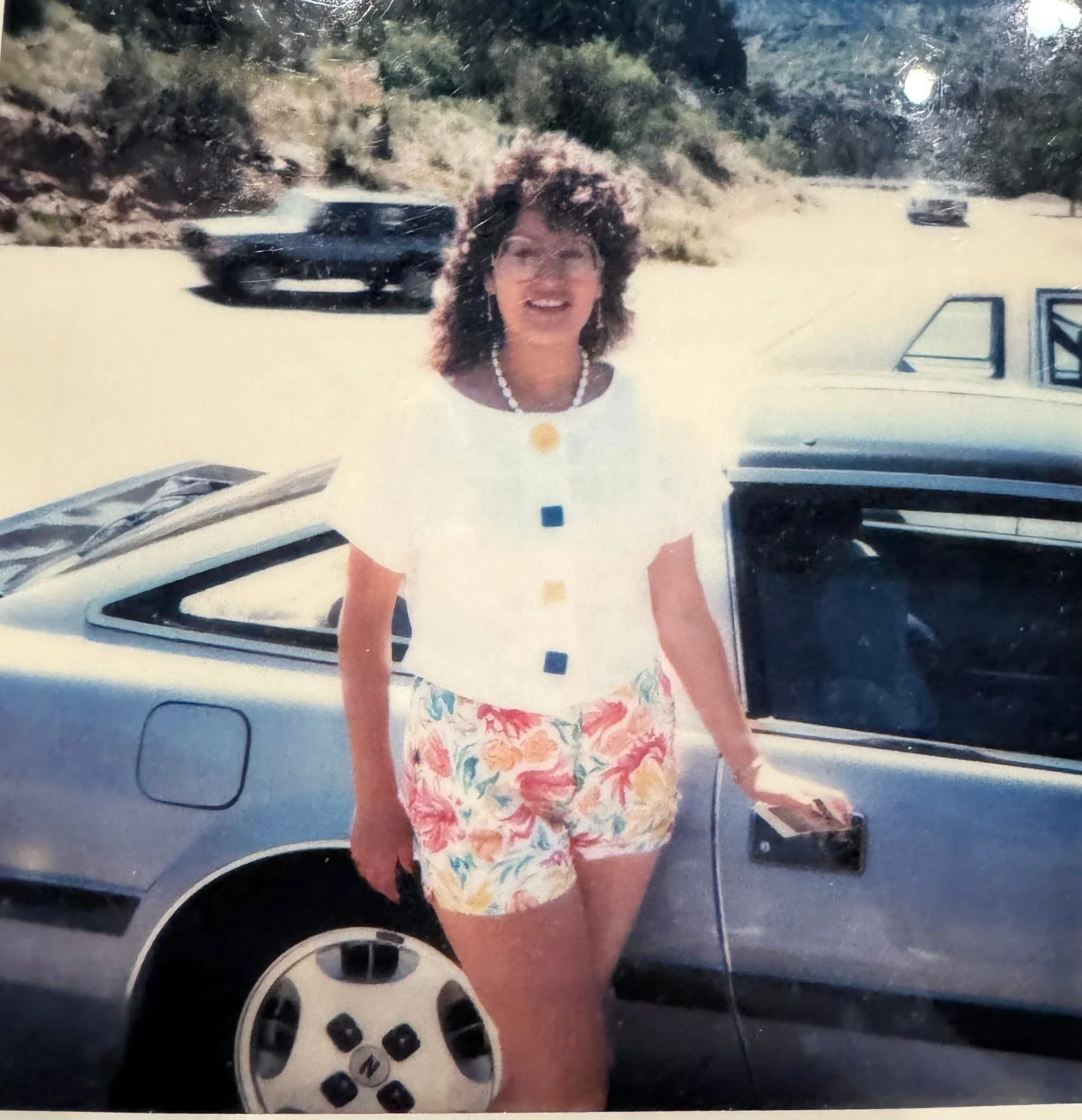 A woman with curly hair, wearing a white top with floral pattern and a pearl necklace, standing outdoors near a white vehicle with open door and smiling.