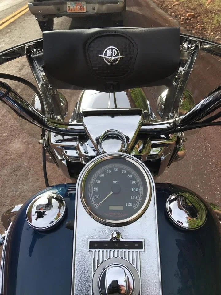 A motorcycle's dashboard with a speedometer, a fuel gauge, and a radio or speaker mounted on the handlebars, with a vehicle ahead on a two-lane road and trees on the side.