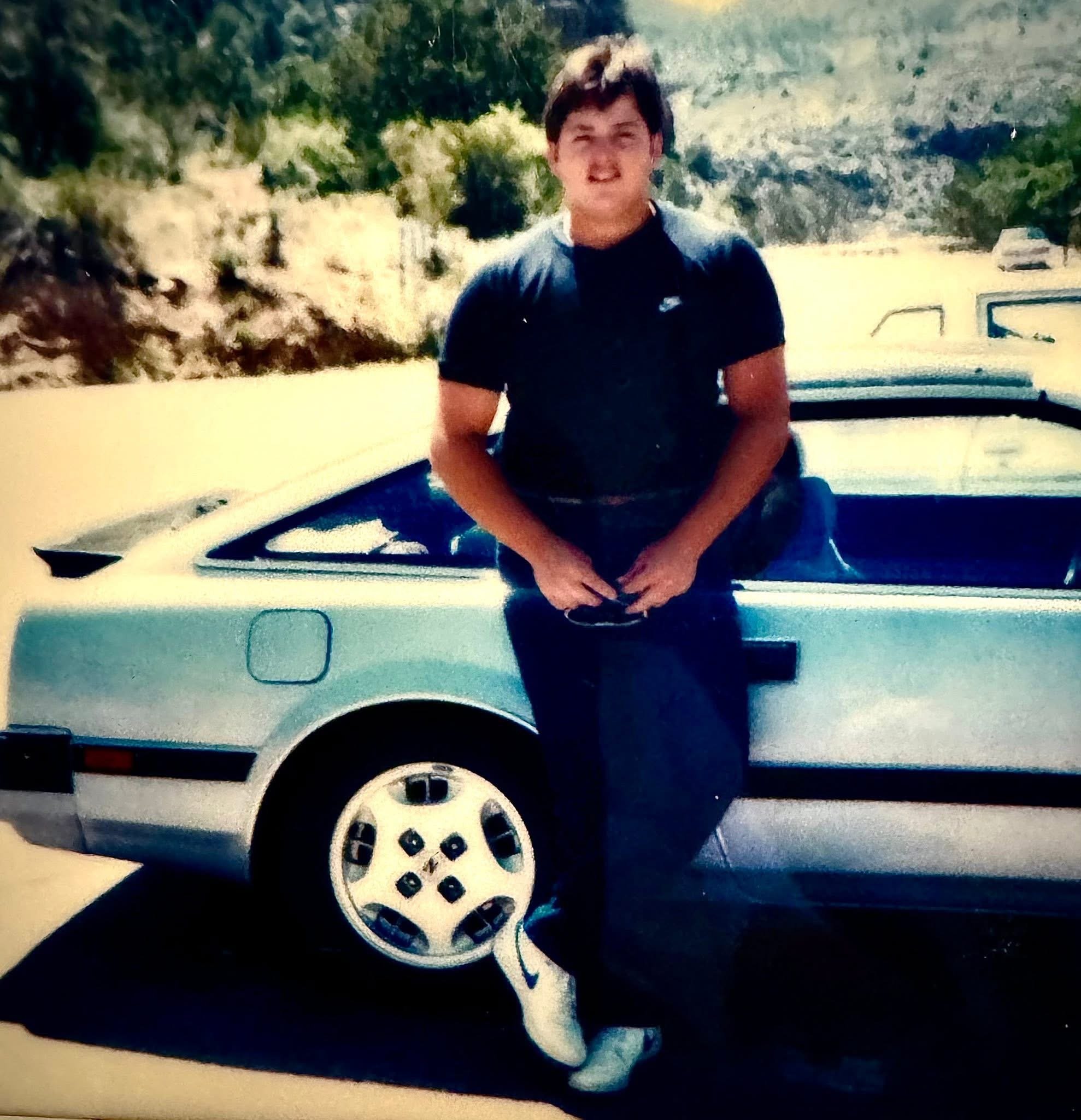 A young person with short brown hair and a slight smile, wearing a black t-shirt and sitting on the hood of a light-colored vehicle, with a background featuring green trees and hills.