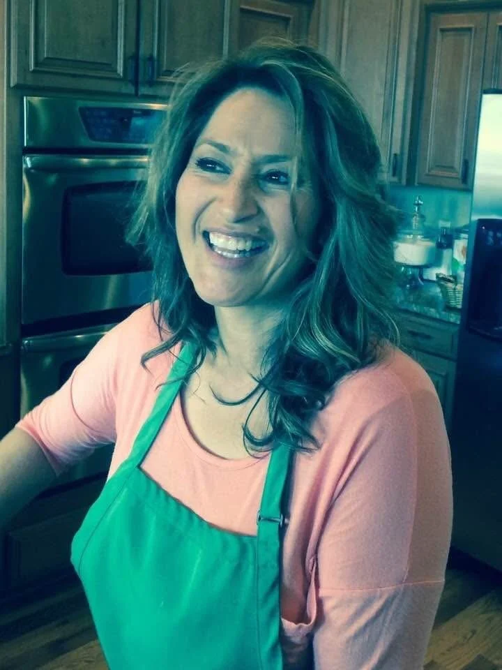 A woman with wavy hair smiling in a kitchen, wearing a pink shirt and a green apron.