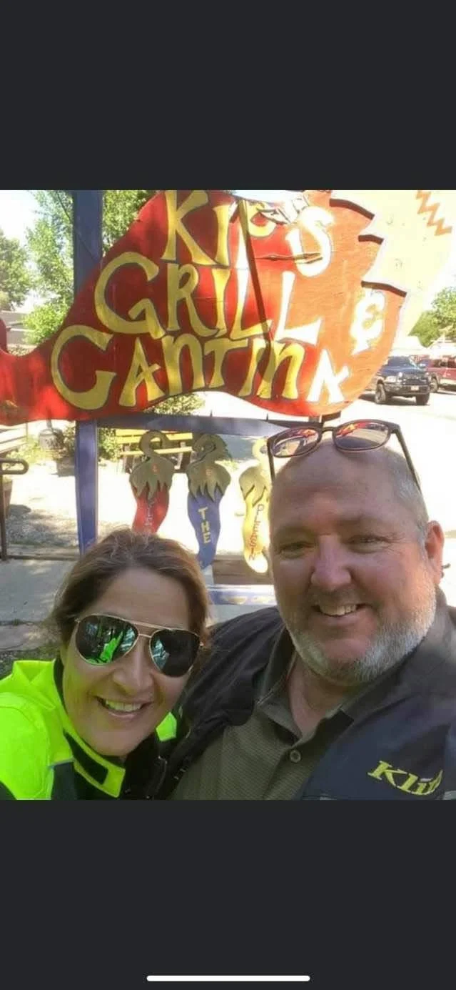 A smiling man and woman taking a selfie in front of a colorful sign that reads "KIDS GRILL & CANTINA" with trees and parked cars in the background.