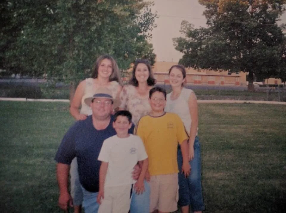A group of six people, three women and three boys, standing outdoors on grass during sunset, smiling for a photo.