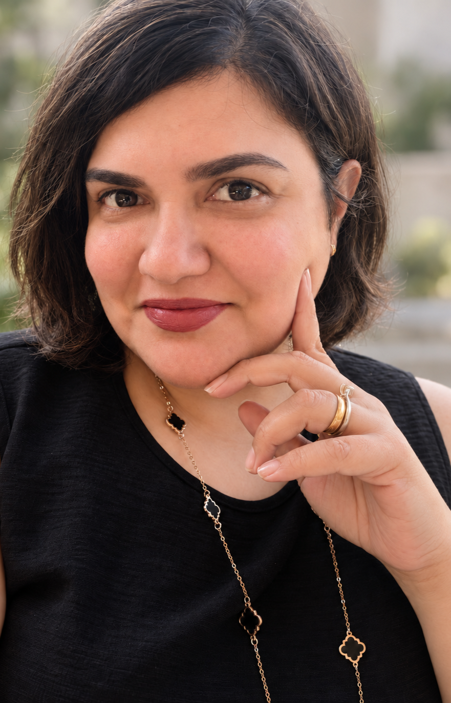 A woman with short dark hair, wearing a black sleeveless top, gold rings, and a gold necklace with black pendant accents, looking at the camera with her hand resting on her chin.