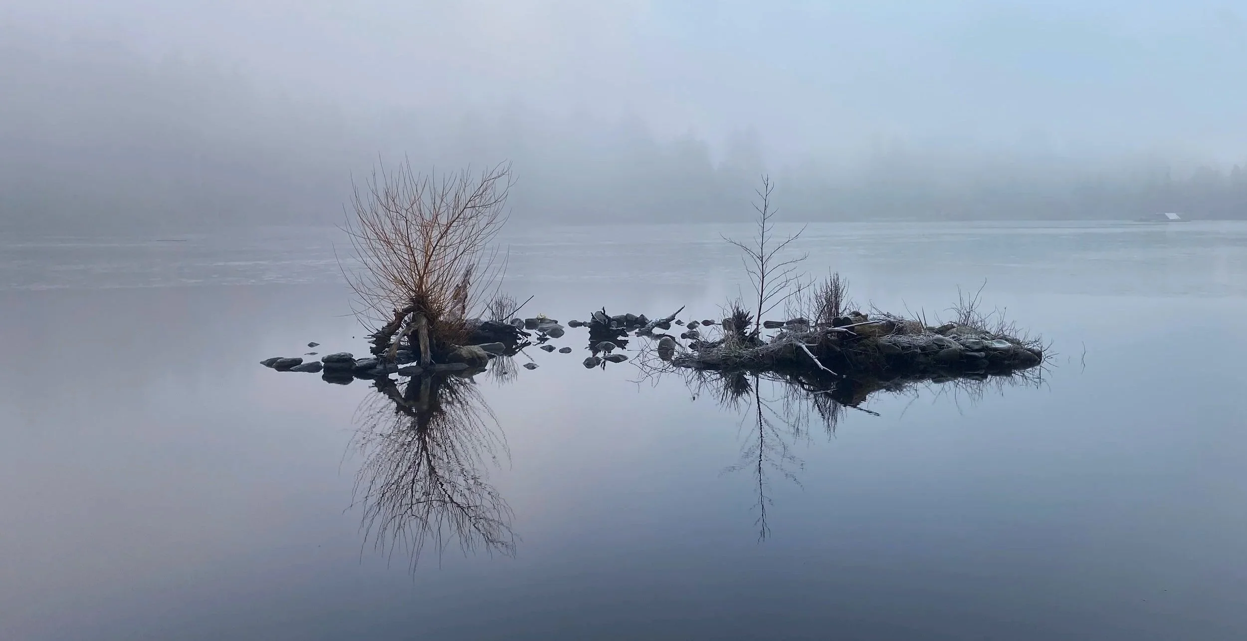 Calm lake with two small tree-covered islands, foggy background, and their reflections in the water.