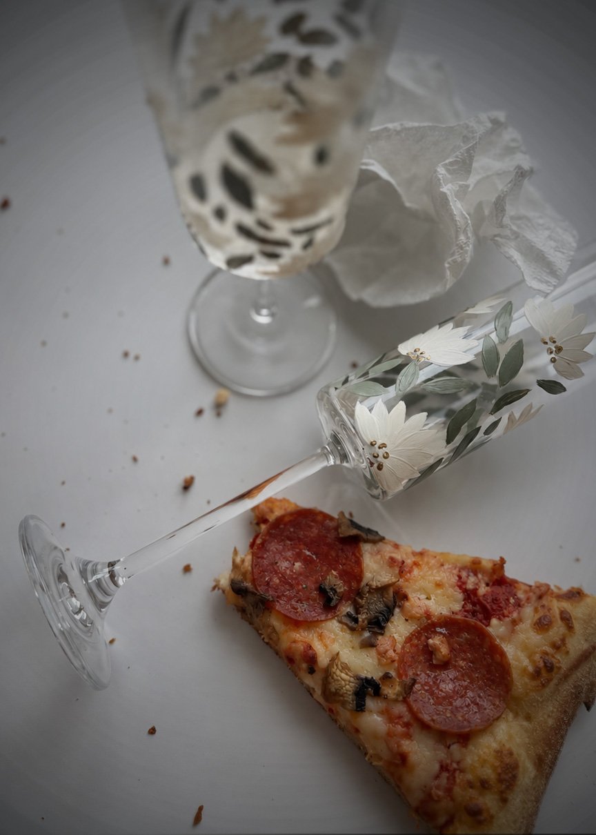 A hand-painted floral champagne flute lying horizontally next to a slice of pepperoni and mushroom pizza on a white surface, with a second flute and crumpled napkin blurred in the background.