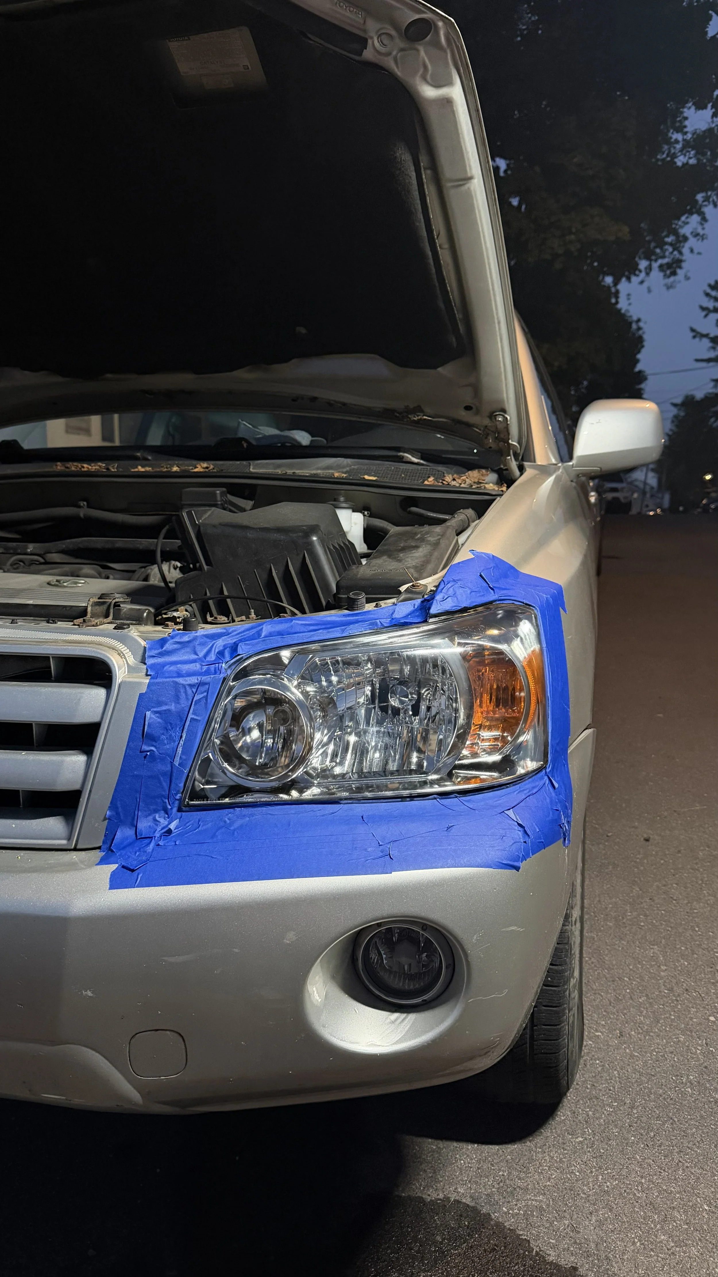Silver car with the front bumper taped off with blue painter's tape, the headlights are visible, and the hood is open showing the engine bay. The car is parked on a street during the evening.