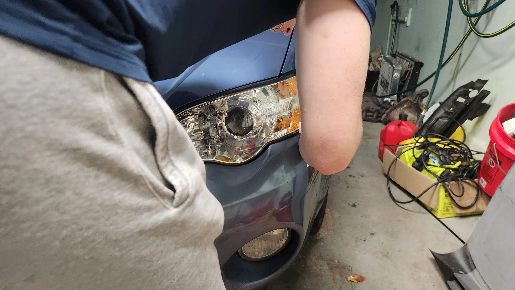 Person working on a car in garage, close-up of front headlight, with tools and equipment nearby.