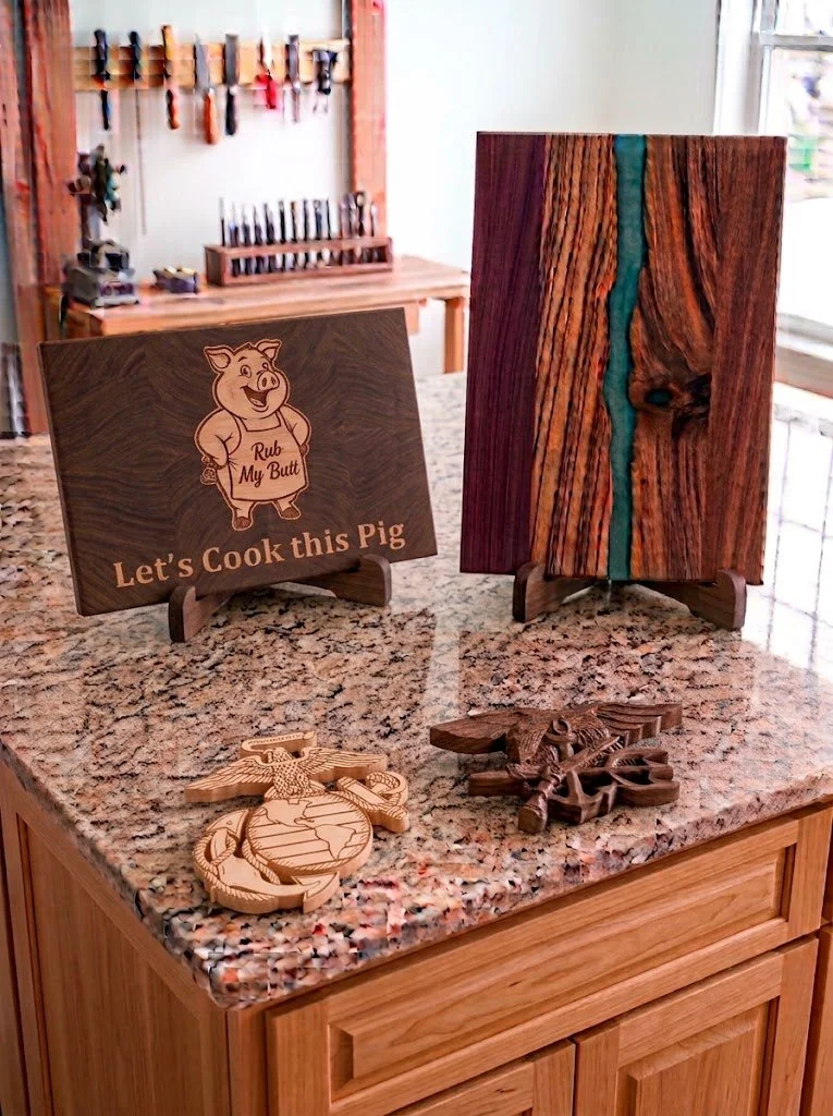 A countertop features a  cutting board with a pig inlay and the text "Let's Cook this Pig" and "Rub My Butt." A river board with turquoise epoxy, Next to a 2d carved EGA in maple, and a 3d carved Navy seal made of walnut