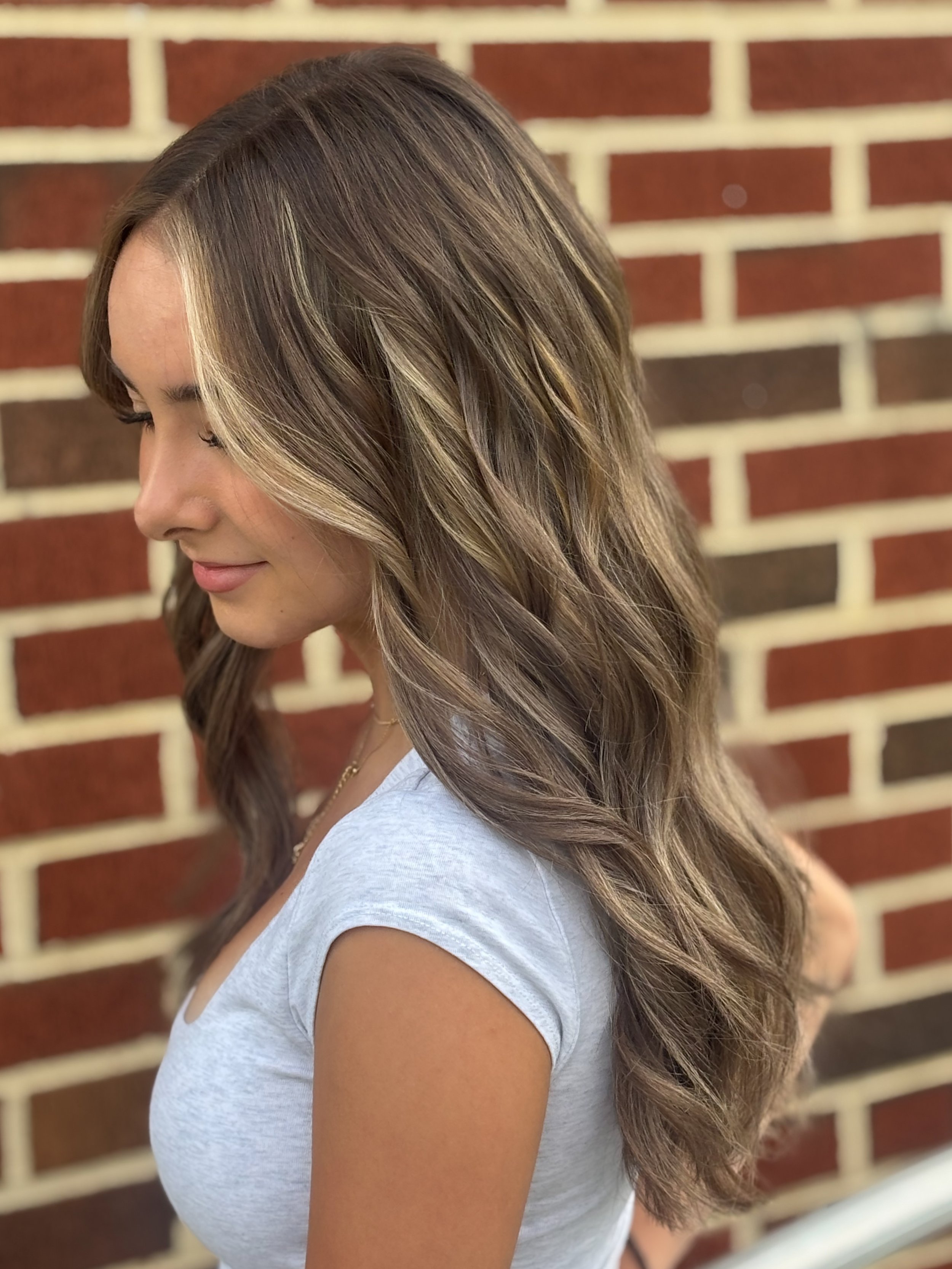 Close-up of a woman with long, wavy, light brown hair with blonde highlights, standing in front of a brick wall.