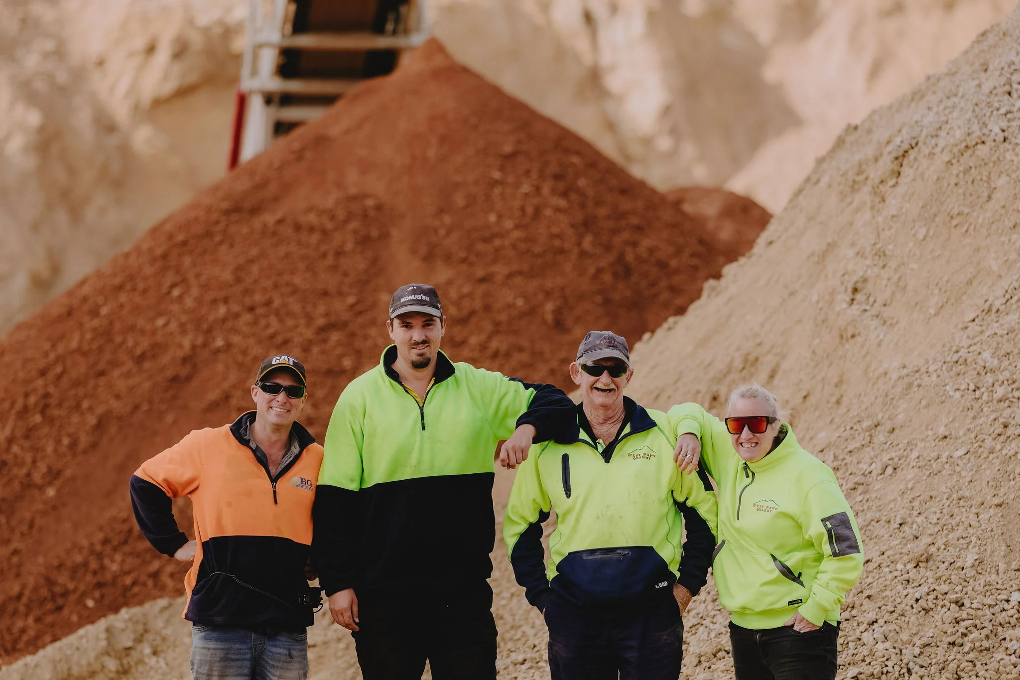 Group of six people standing in front of large rocks at a quarry, smiling and wearing high-visibility jackets and safety glasses.