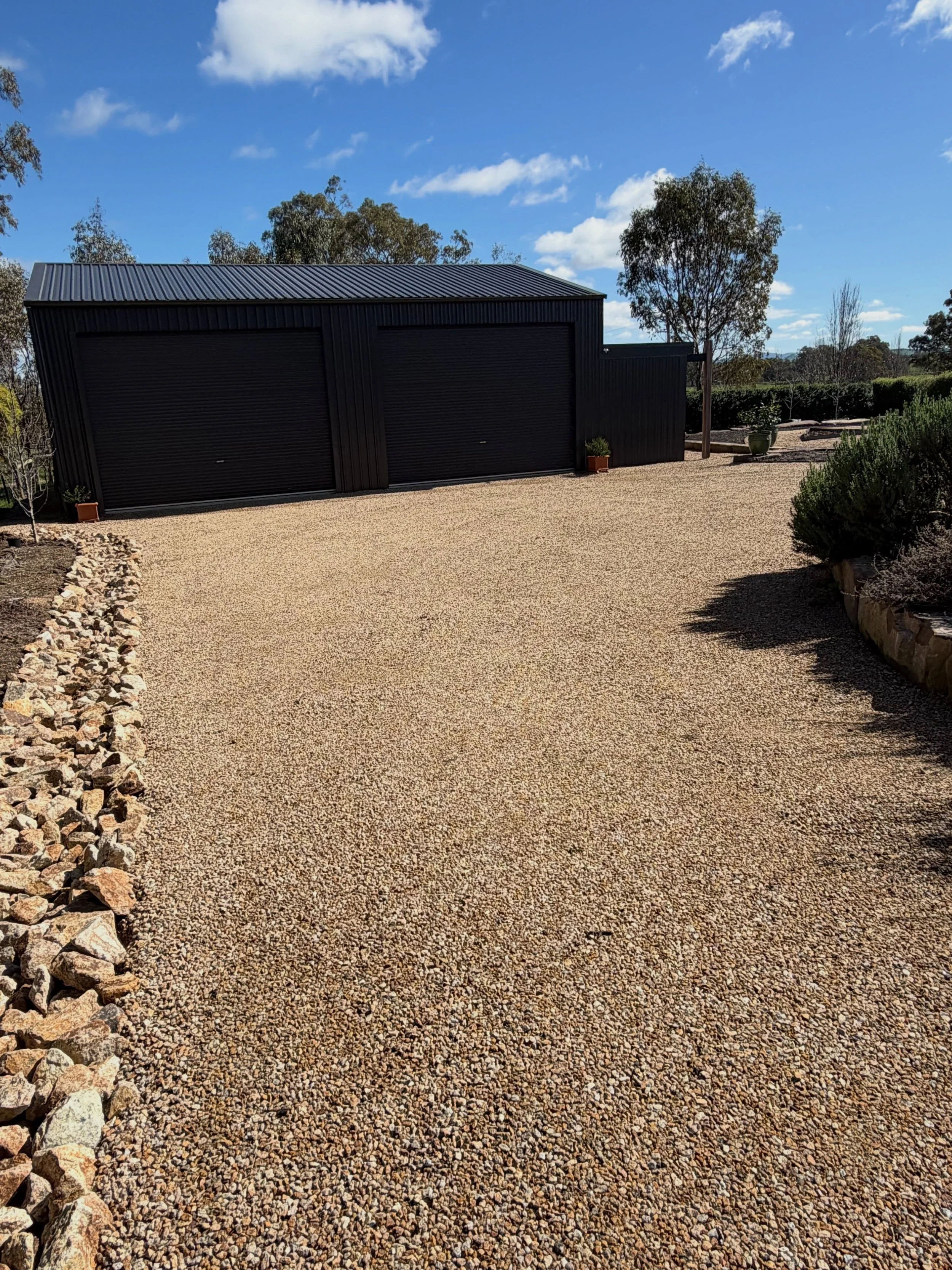 A black building with two closed roller doors, surrounded by a gravel driveway, potted plants, and trees under a blue sky with some clouds.