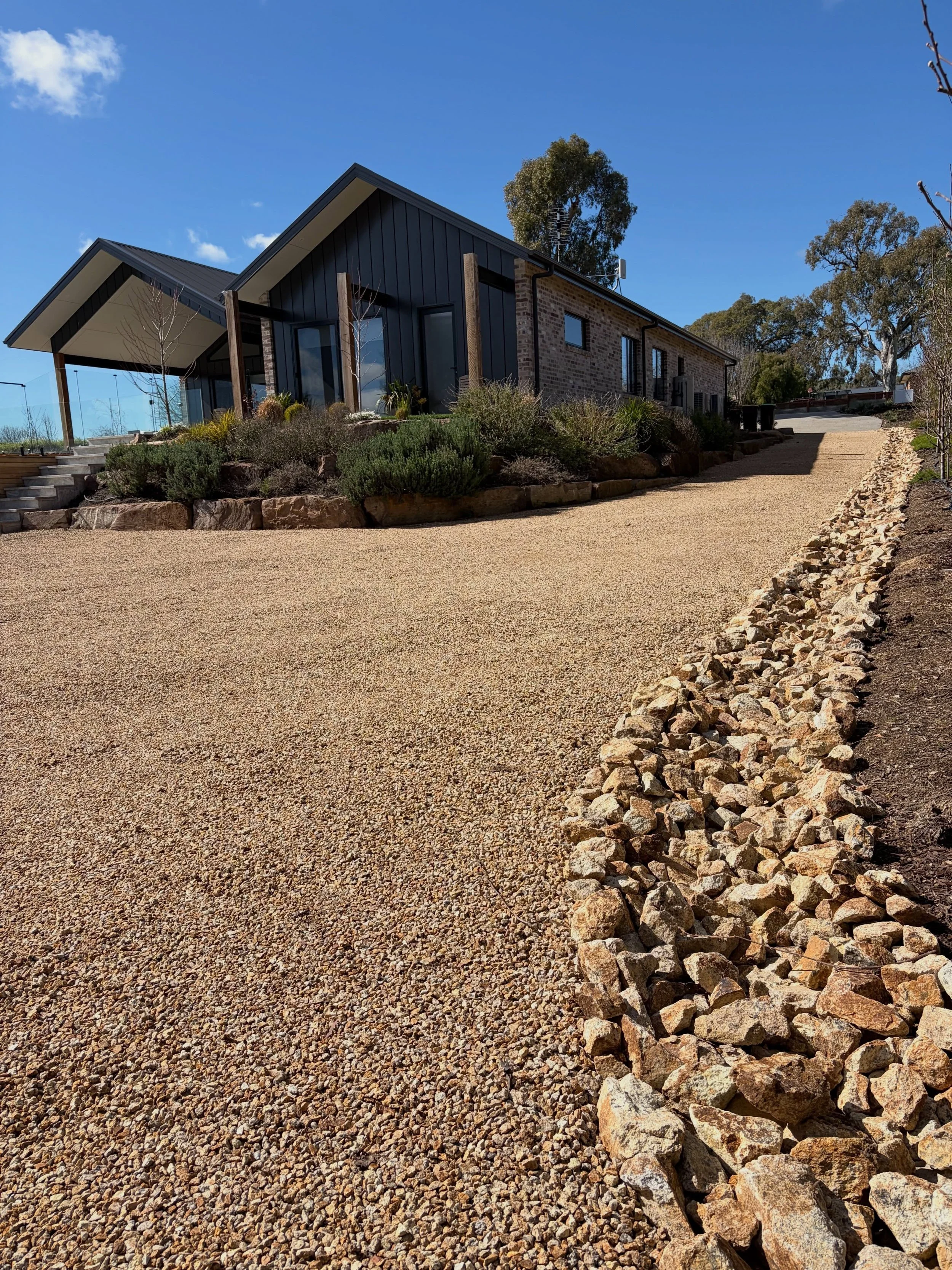 A modern house with a gravel driveway, landscaped garden, and a hillside. The house has a dark metal roof, large glass windows, and a brick and metal exterior. There are tall trees and a clear blue sky.