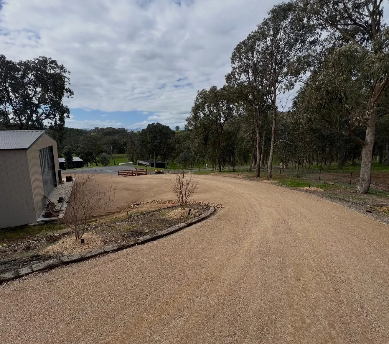 An unpaved gravel driveway winding through a rural landscape with trees and a shed on the left. A small tree is in the foreground, and there are trees and hills in the background under a partly cloudy sky.