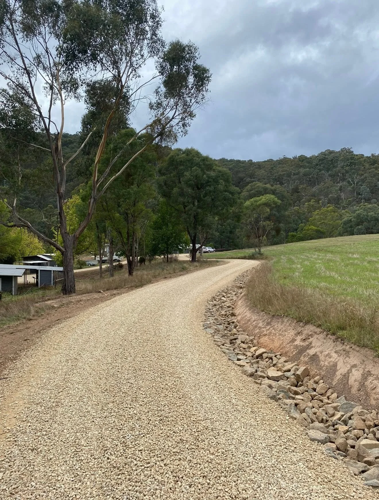 A gravel road winding through a rural area with trees on the left and a grassy field on the right, with hills and cloudy skies in the background.