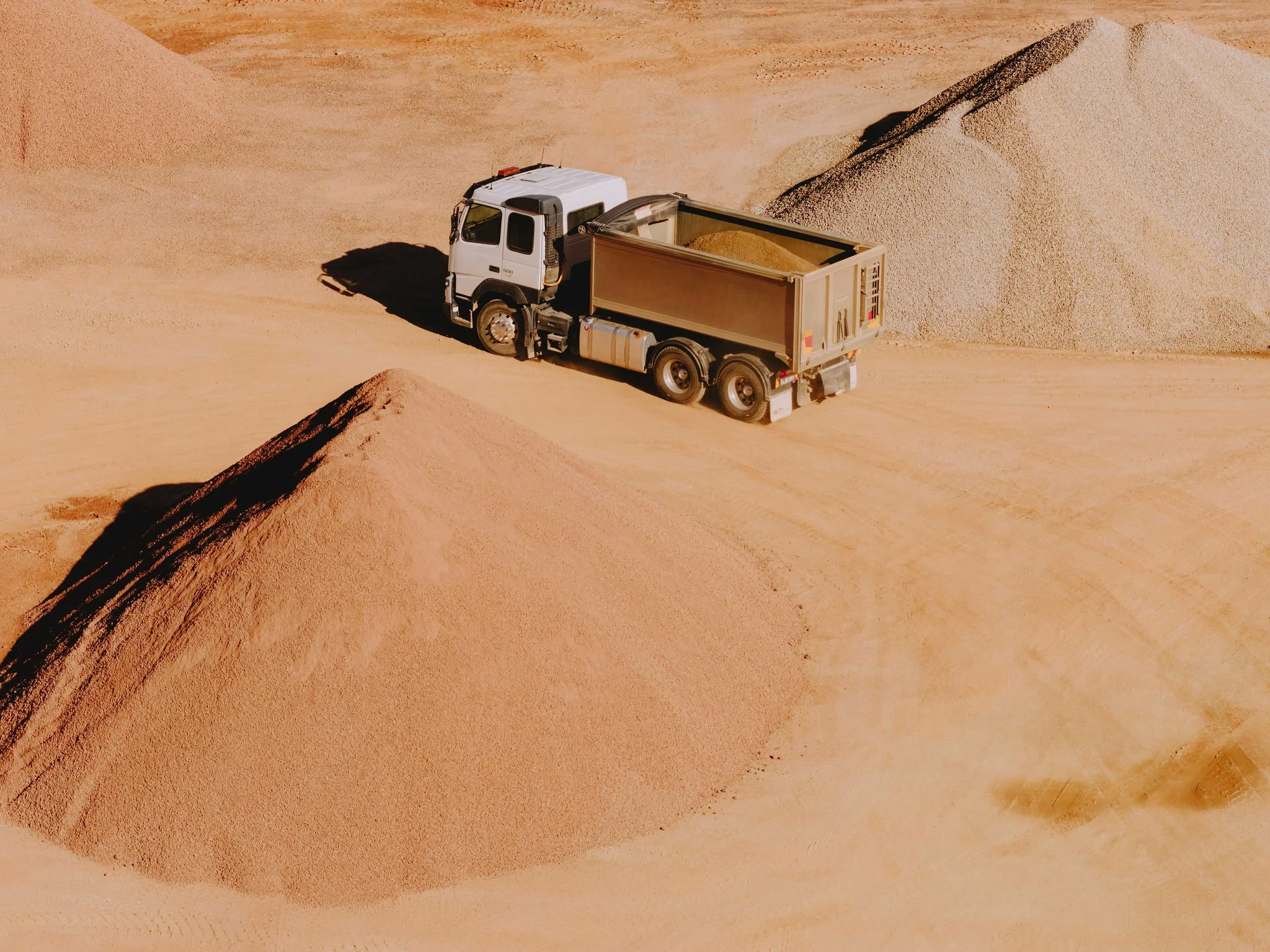 A white truck with an open bed filled with sand, driving through a desert landscape with sand dunes.