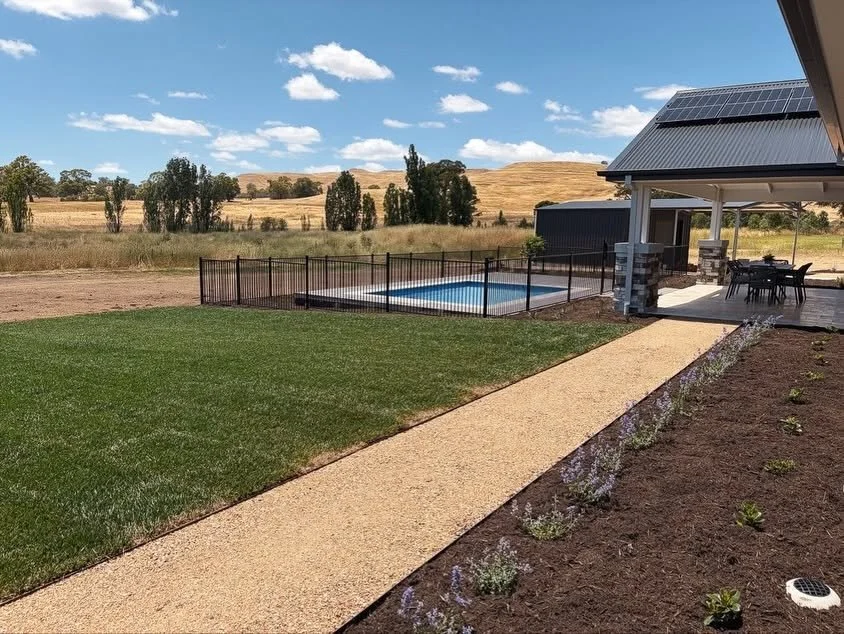 View of a backyard with a green lawn, a small pool enclosed by a black fence, a concrete walkway, and a covered patio area with outdoor furniture, set against a rural landscape with grassy hills and a partly cloudy sky.