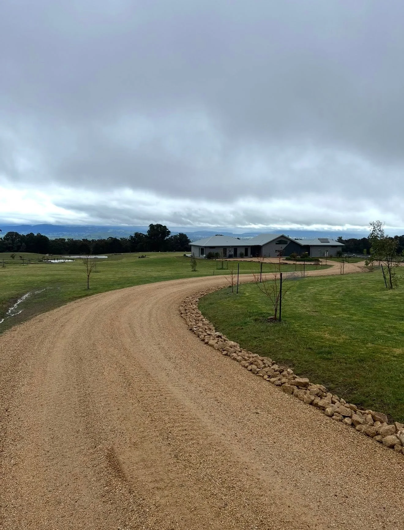 A gravel driveway winding through a grassy area with young trees, leading to a modern house under a cloudy sky.