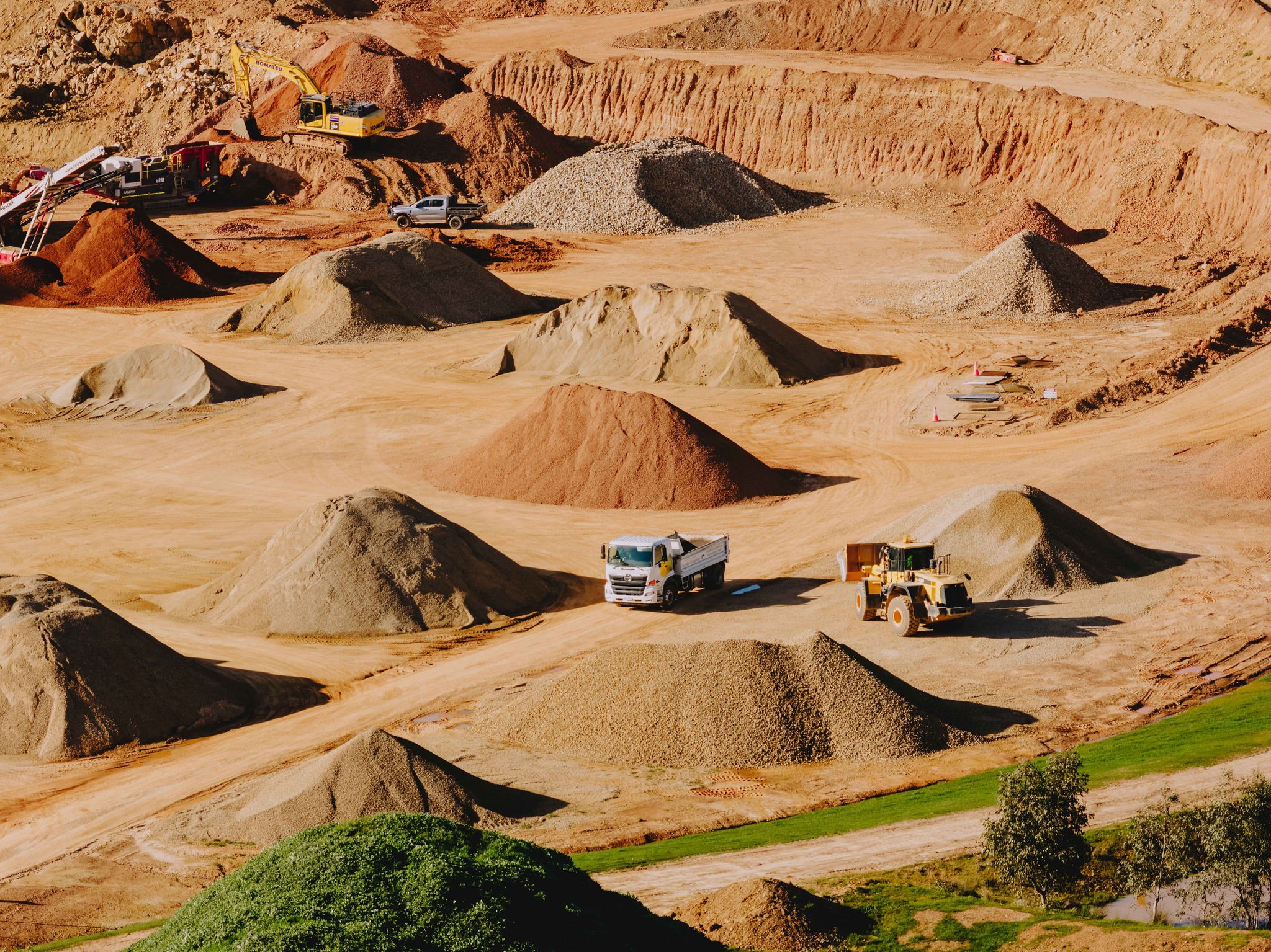 Construction site on a hillside with equipment and piles of dirt, rocks, and gravel. Some green grass and trees are visible in the foreground.