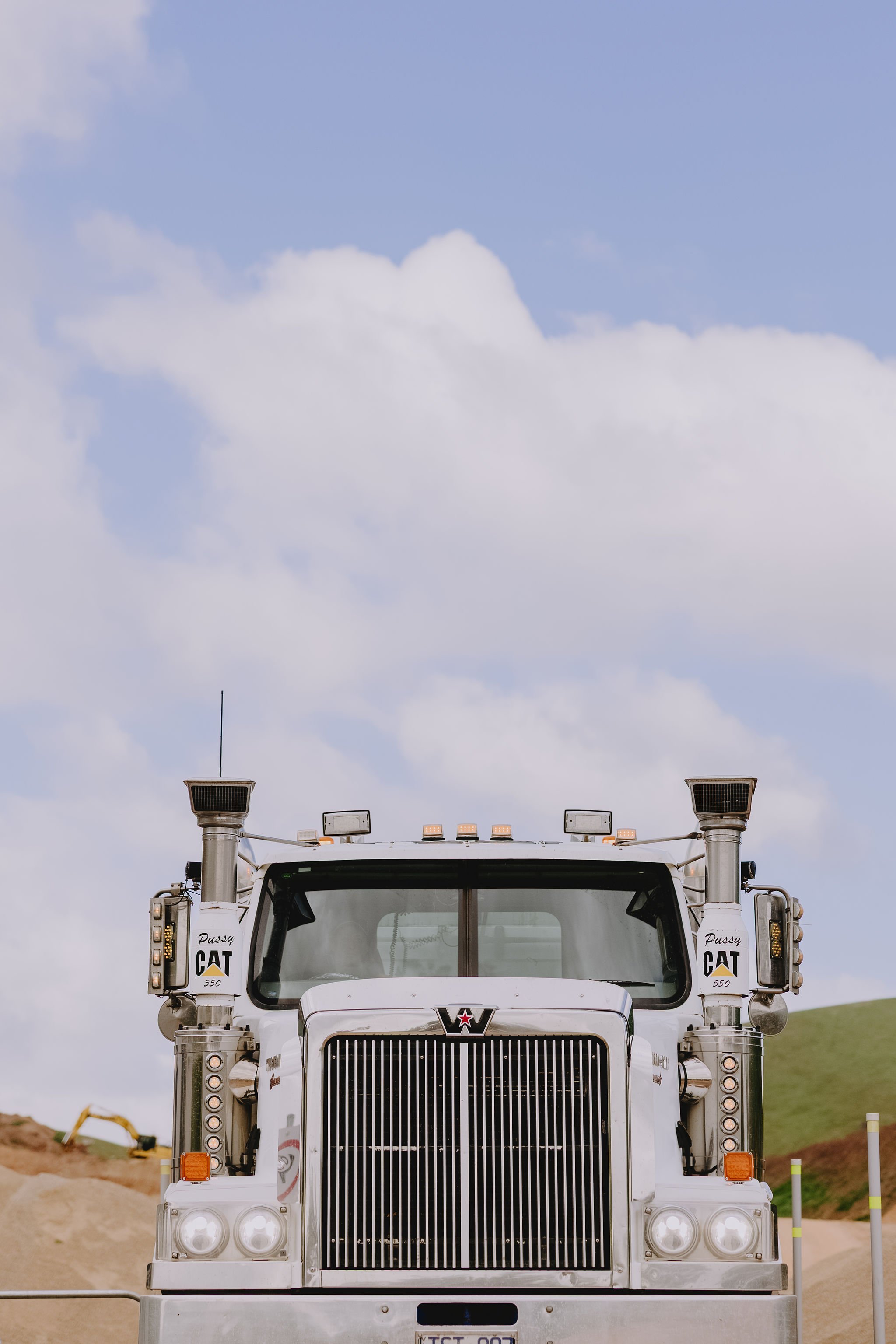 Front view of a large white semi-truck with a black grille, multiple lights, and two side mirrors, parked outdoors under a partly cloudy sky.
